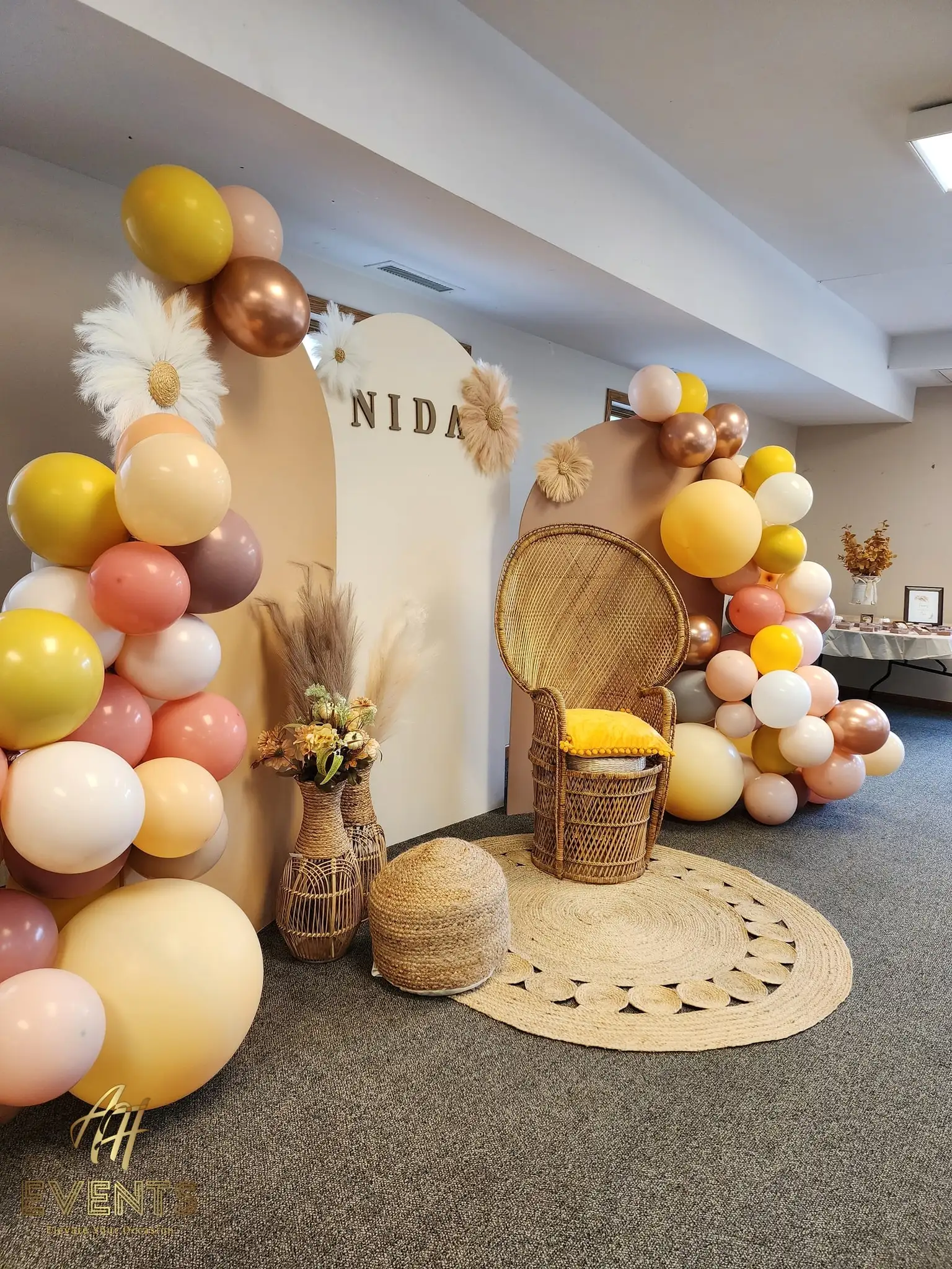 Boho baby shower backdrop with arches reading 'Nida', a mustard and rust balloon garland and a peacock wicker chair
