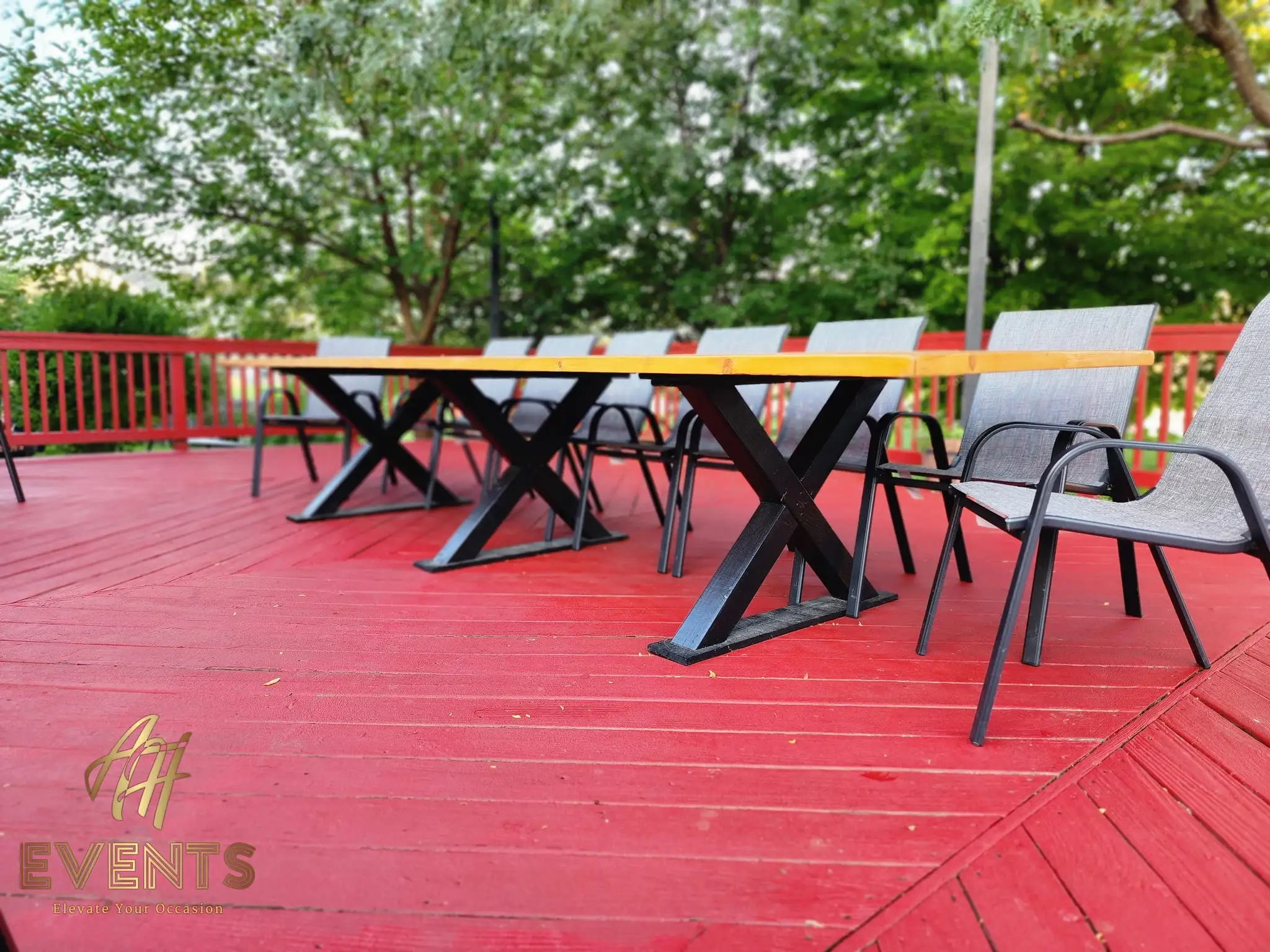 Handcrafted wooden farm table with black X-frame legs and gray patio chairs on a red deck, angled view among trees