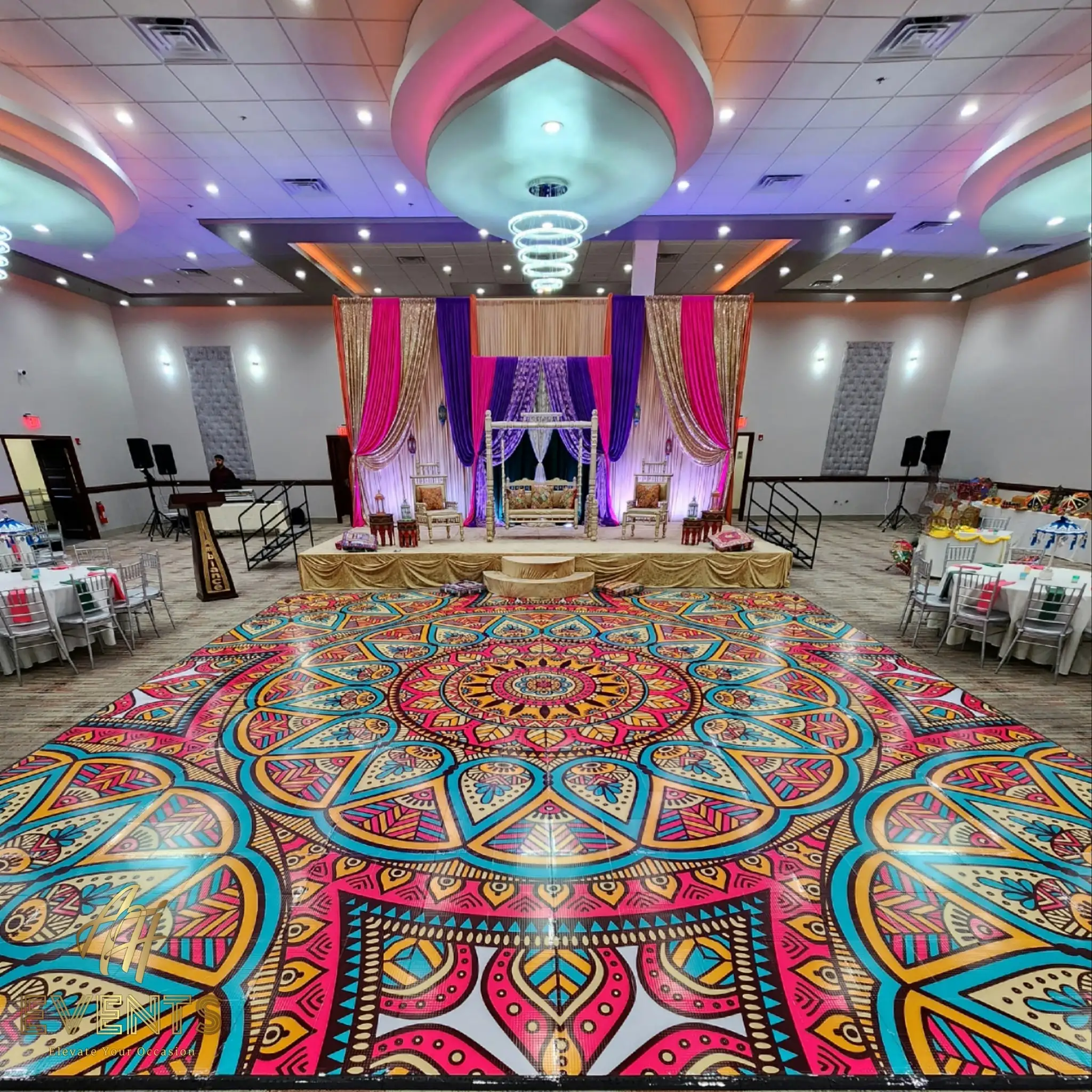 Large circular mandala dance floor in pink, teal and gold filling a ballroom, with a pink and purple draped stage