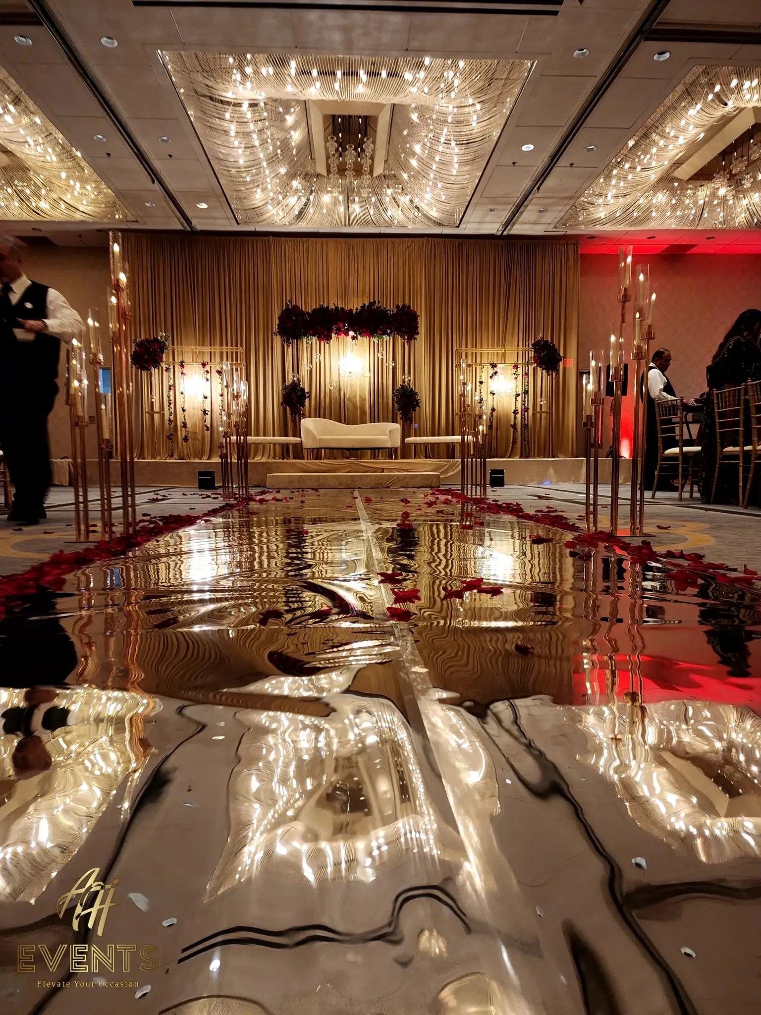 Gold mirror-finish vinyl aisle reflecting ballroom chandeliers, leading to a draped stage with a white sofa and red rose accents