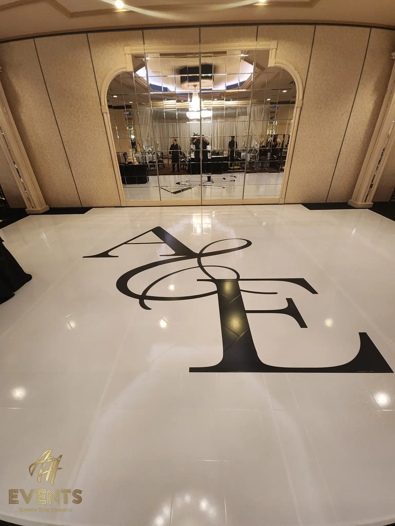 Glossy white dance floor with a large black 'A&E' serif monogram, reflected in the mirrored ballroom wall