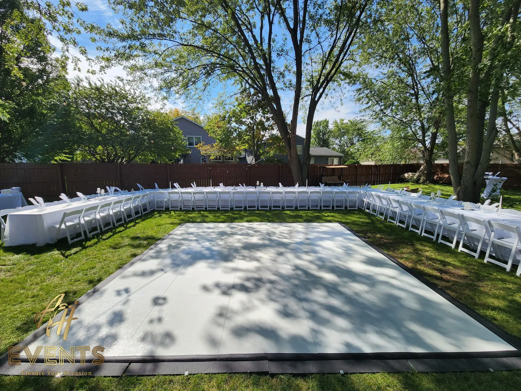 White dance floor on a backyard lawn surrounded by white banquet tables and chairs, framed by trees under a blue sky