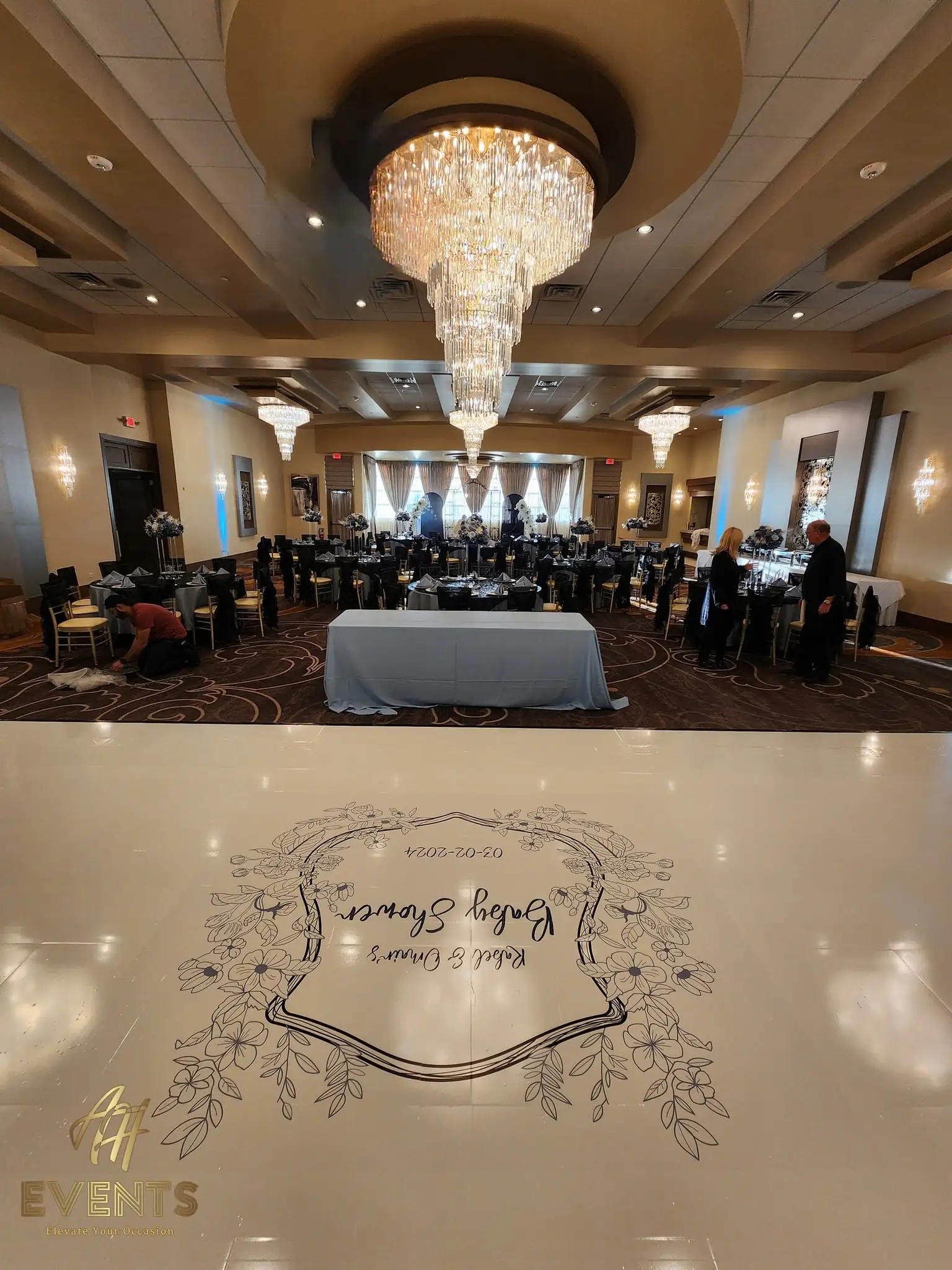 Glossy cream dance floor printed with a 'Rabel & Omar's Baby Shower, 03-02-2024' floral-crest decal, a head table and black-chair tables behind