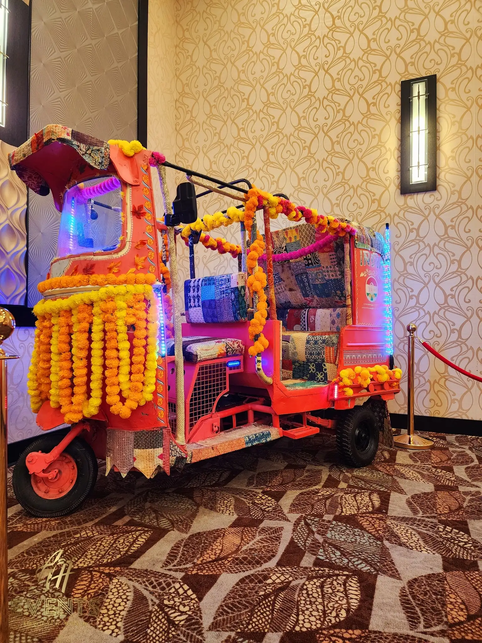 Auto rickshaw decorated with marigold garlands and a glowing LED interior, displayed indoors behind a rope barrier