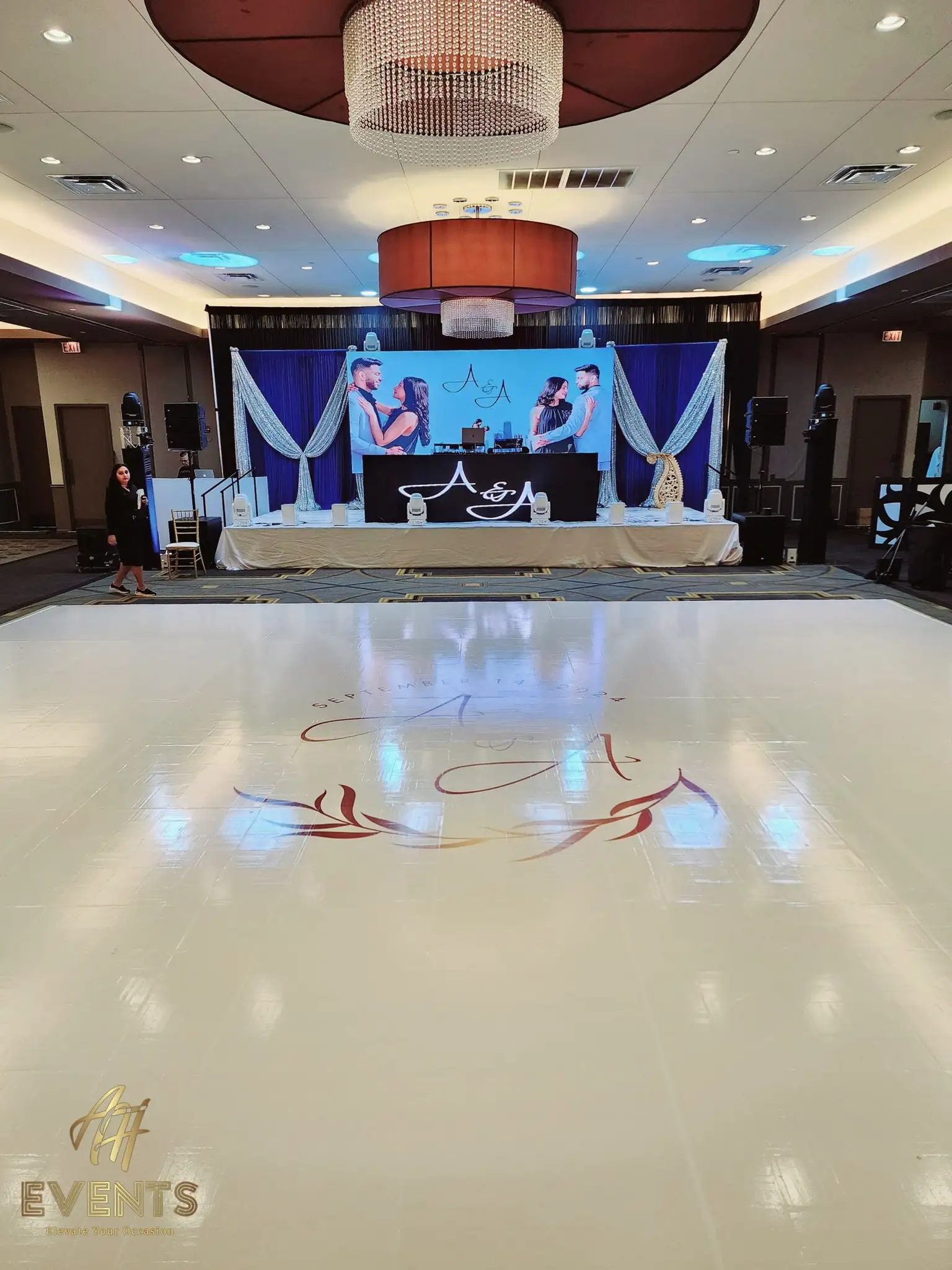 Glossy white dance floor printed with an 'A & A' monogram and foliage wreath, facing a navy stage with the couple's portrait screen and a head table
