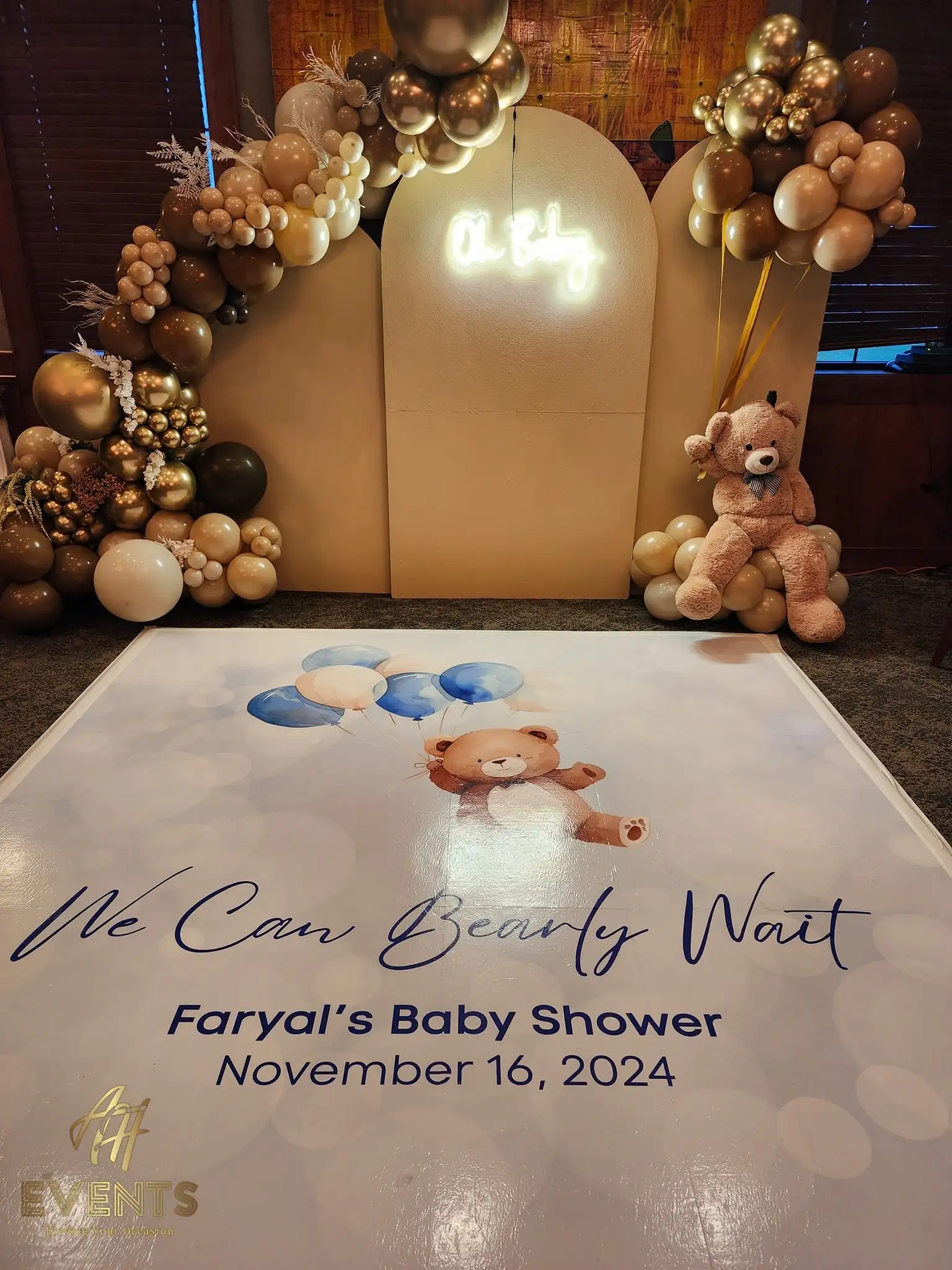 Bear-themed baby shower with tan arch backdrops, a brown-and-gold balloon garland, teddy bears, a neon 'Oh Baby' sign and a printed floor decal reading 'We Can Bearly Wait — Faryal's Baby Shower'