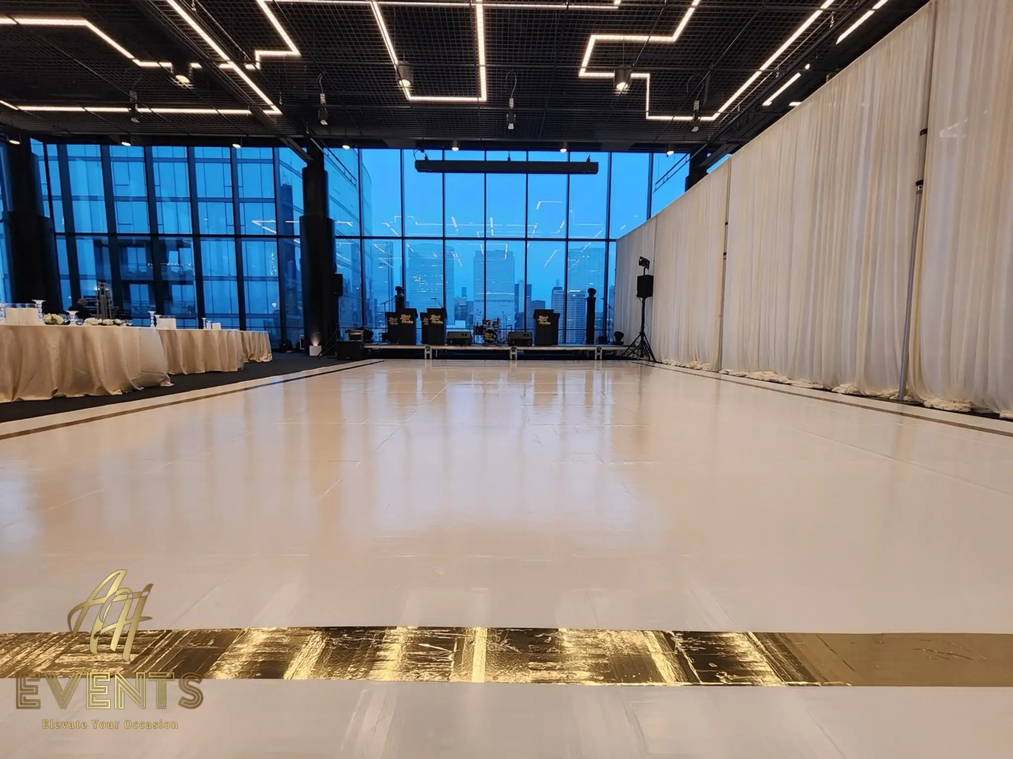 Glossy white dance floor with a gold mirror-stripe border in a loft venue, a band stage and city skyline windows behind
