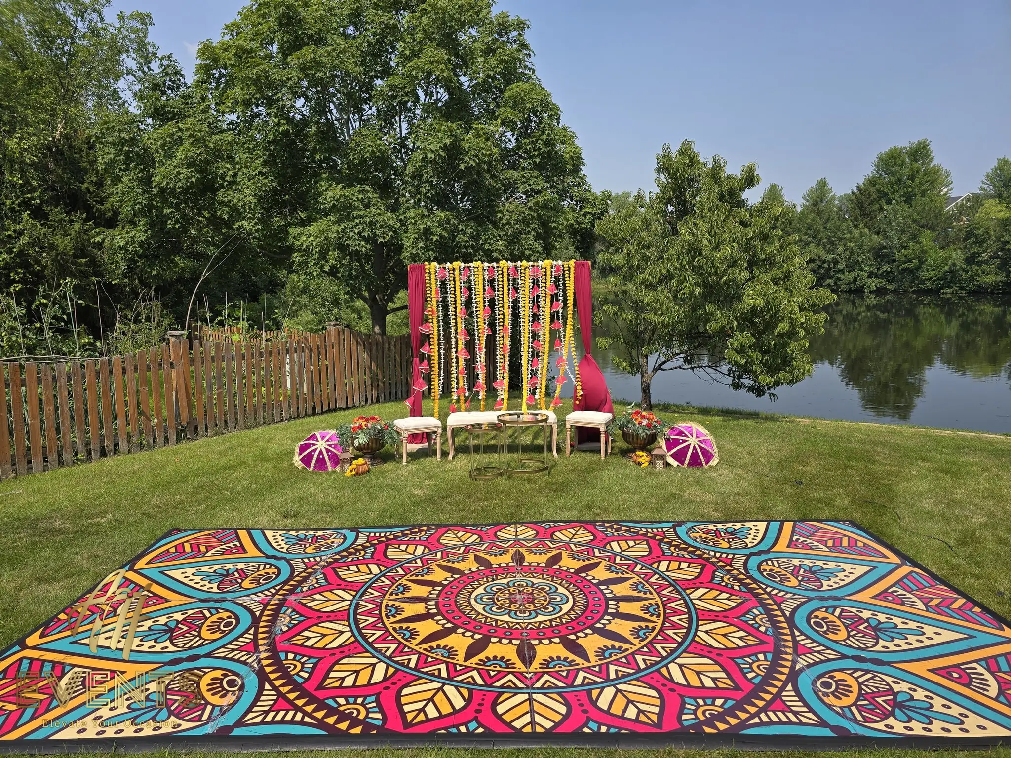 Mandala floor set on a lawn beside a pond, with a magenta floral backdrop and decorative parasols