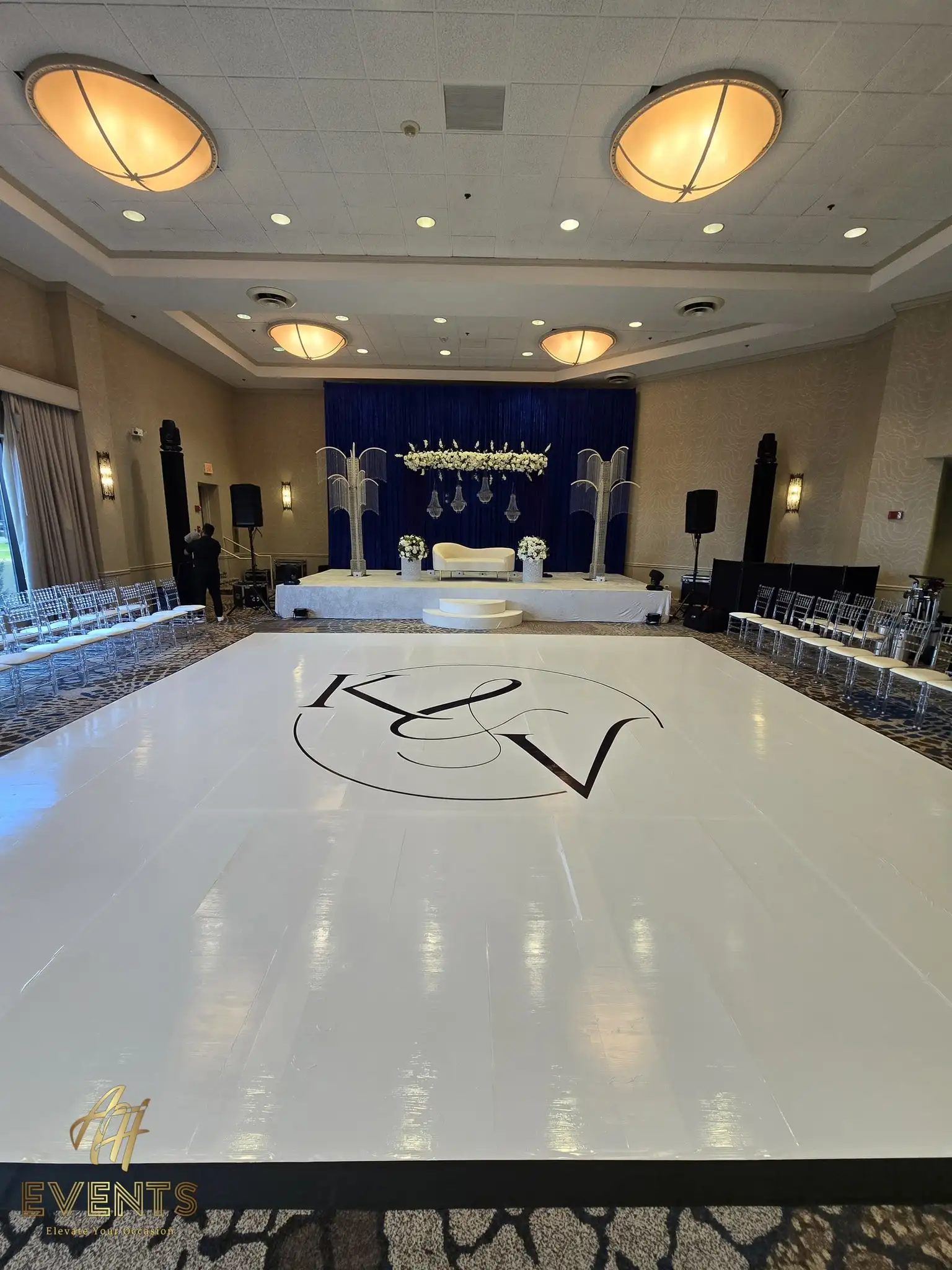 Glossy white dance floor with a 'K&V' monogram facing a navy stage flanked by crystal palm columns and a floral garland
