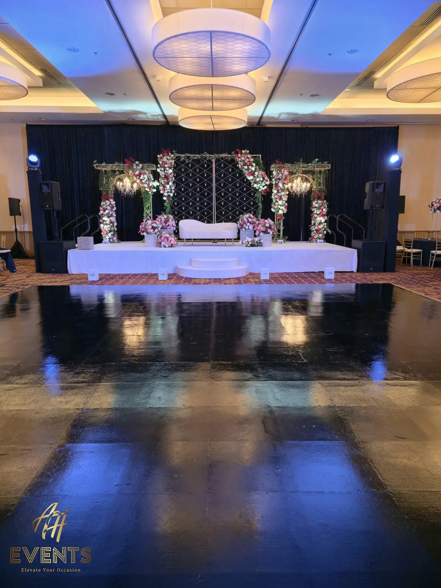 Glossy black dance floor reflecting a white floral mandap stage and drum chandeliers in a ballroom
