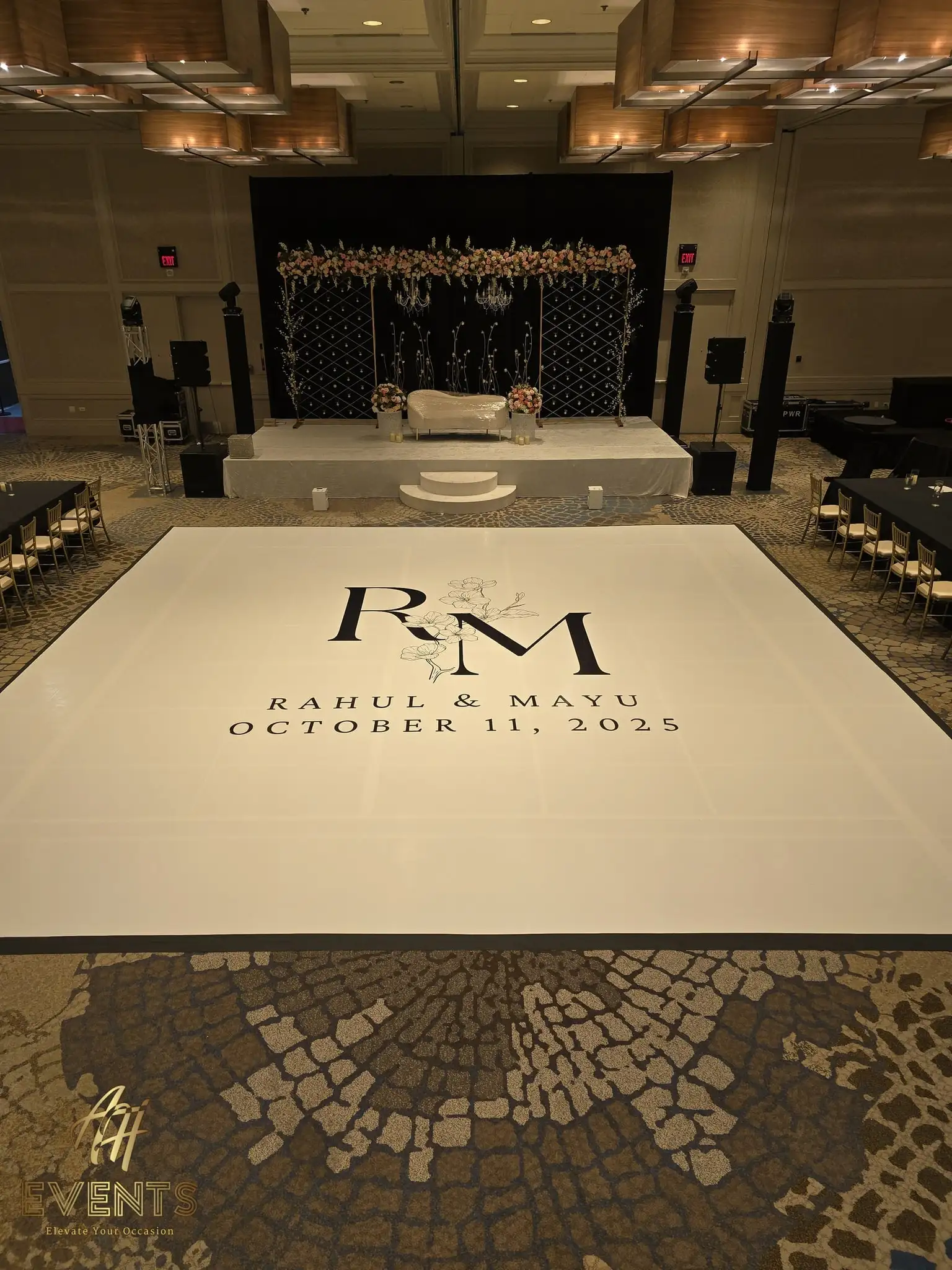 Glossy white dance floor printed with an 'R M — Rahul & Mayu, October 11, 2025' magnolia monogram, a black lattice mandap stage behind