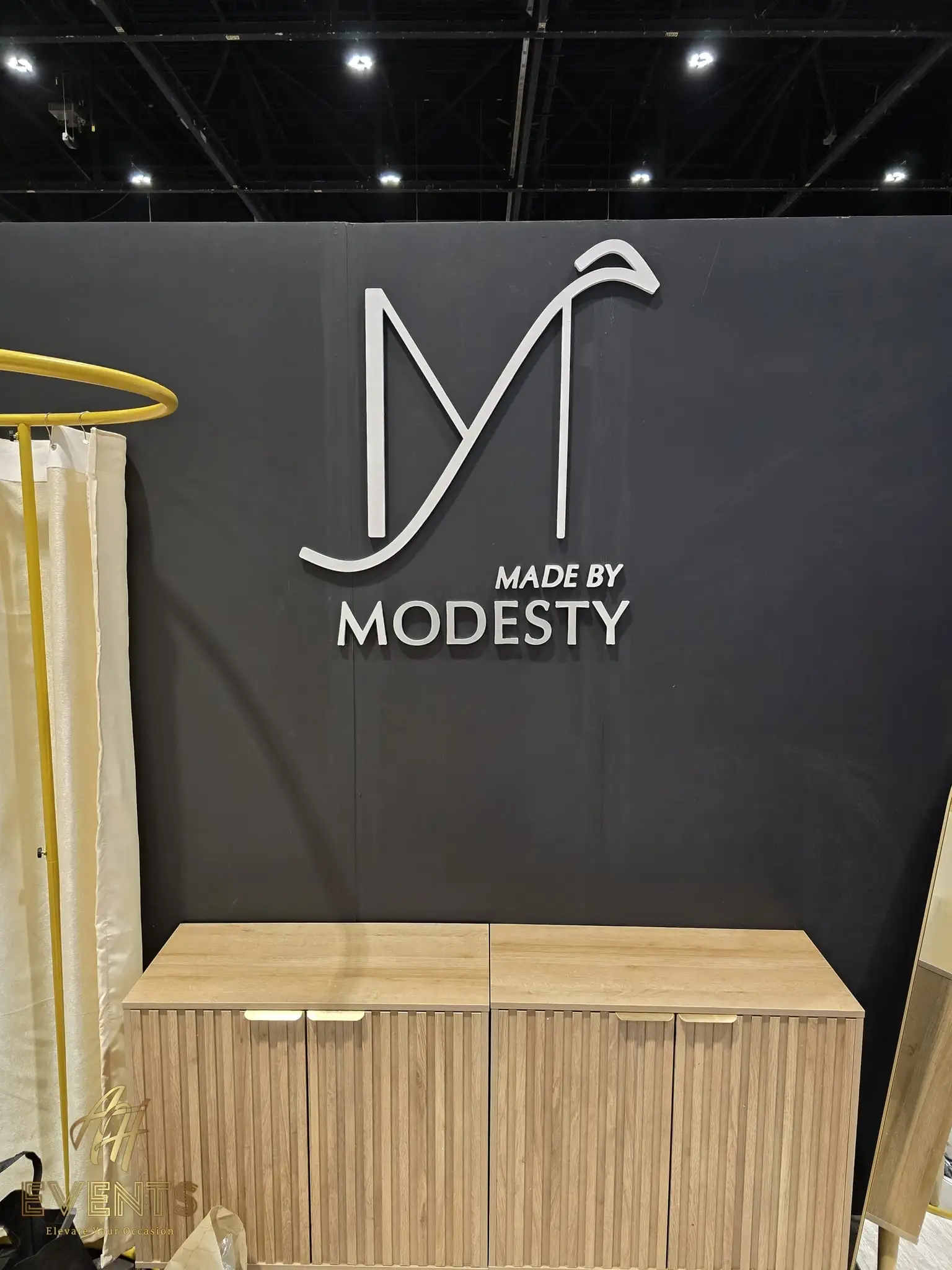 Made by Modesty convention booth with a white 'M' logo on a dark wall above a fabricated wood cabinet