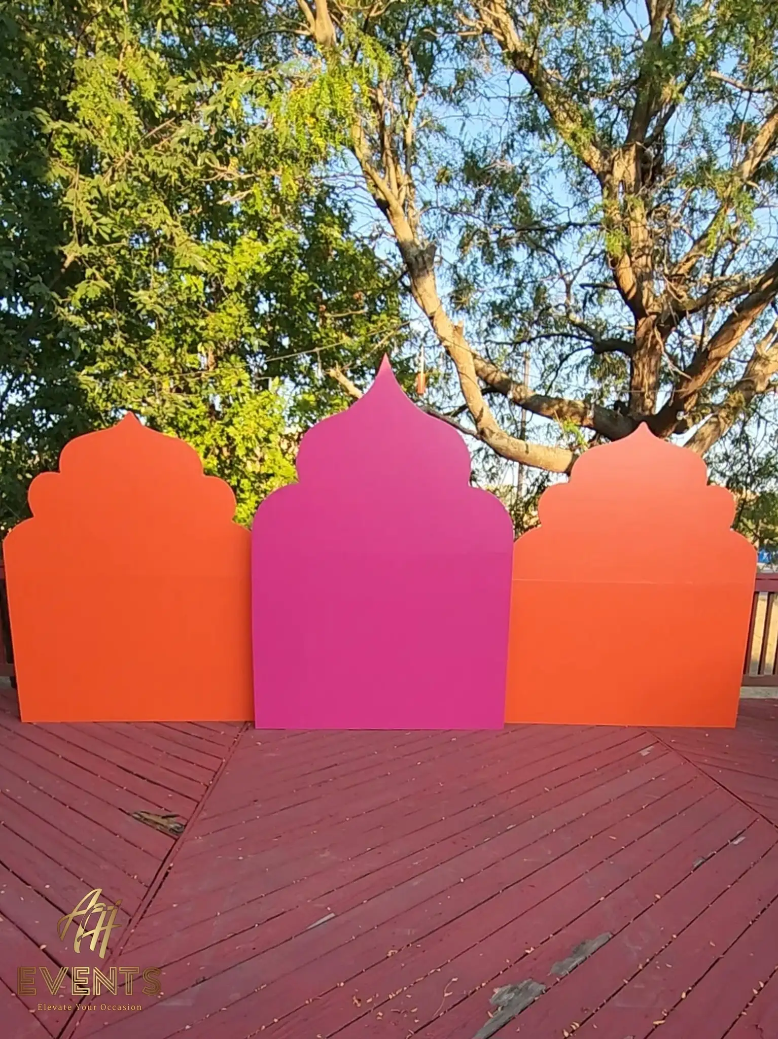 Three Mughal-arch backdrop panels in orange, magenta and orange standing on a red deck among bare trees