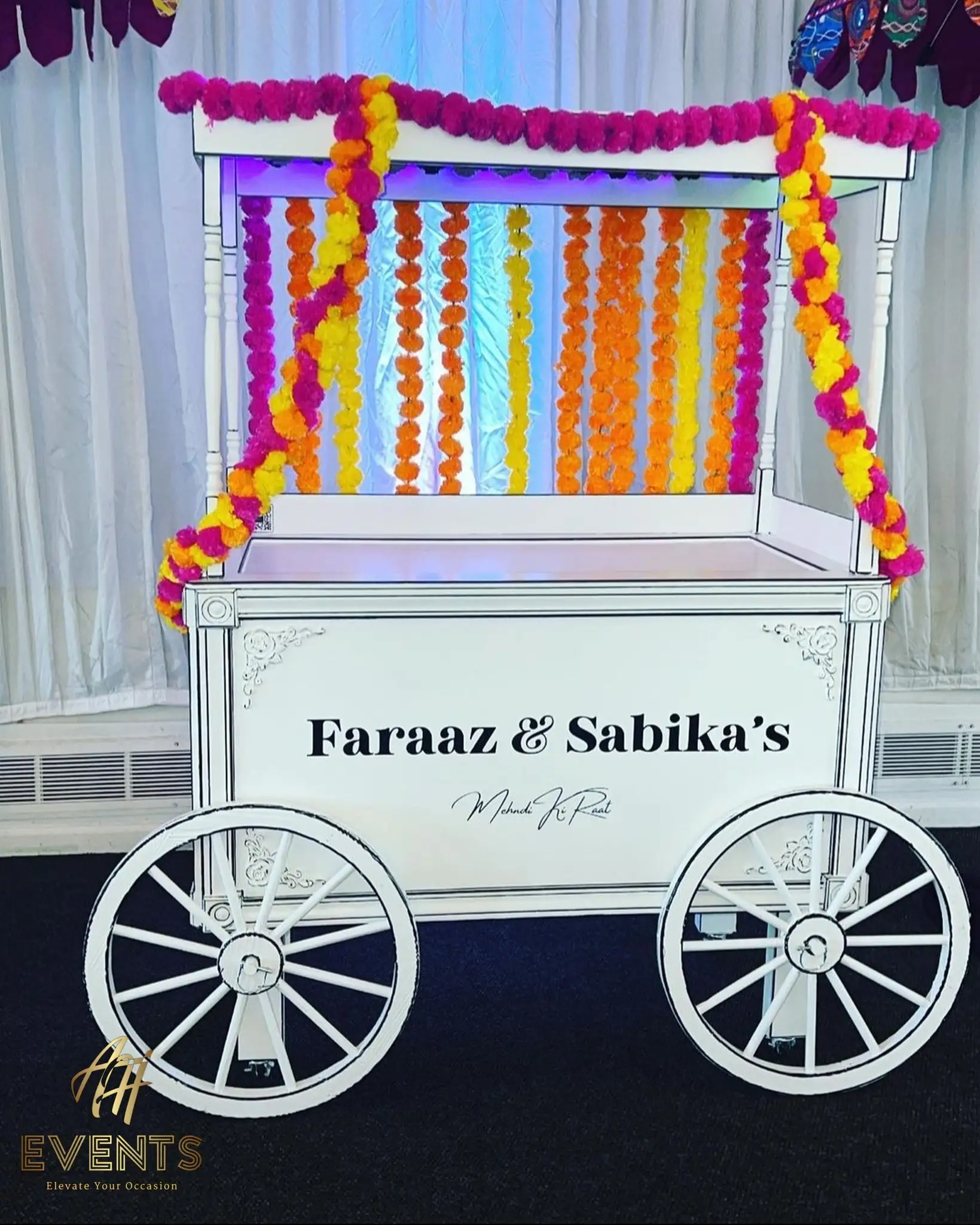 White wooden favor cart with pink and orange marigold garlands and 'Faraaz & Sabika's' lettering on the front