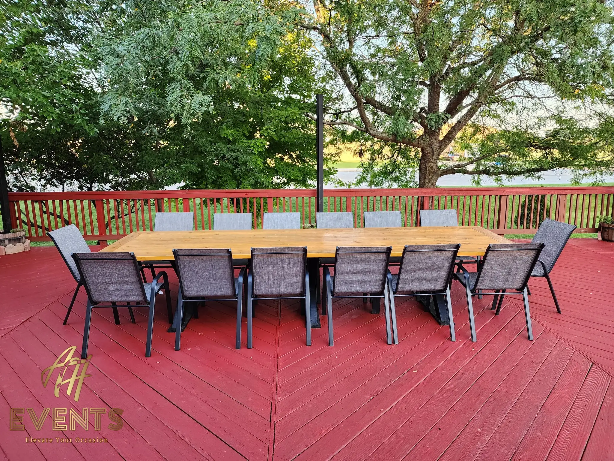 Long wooden farm table set with ten gray patio chairs on a red deck, trees behind at sunset