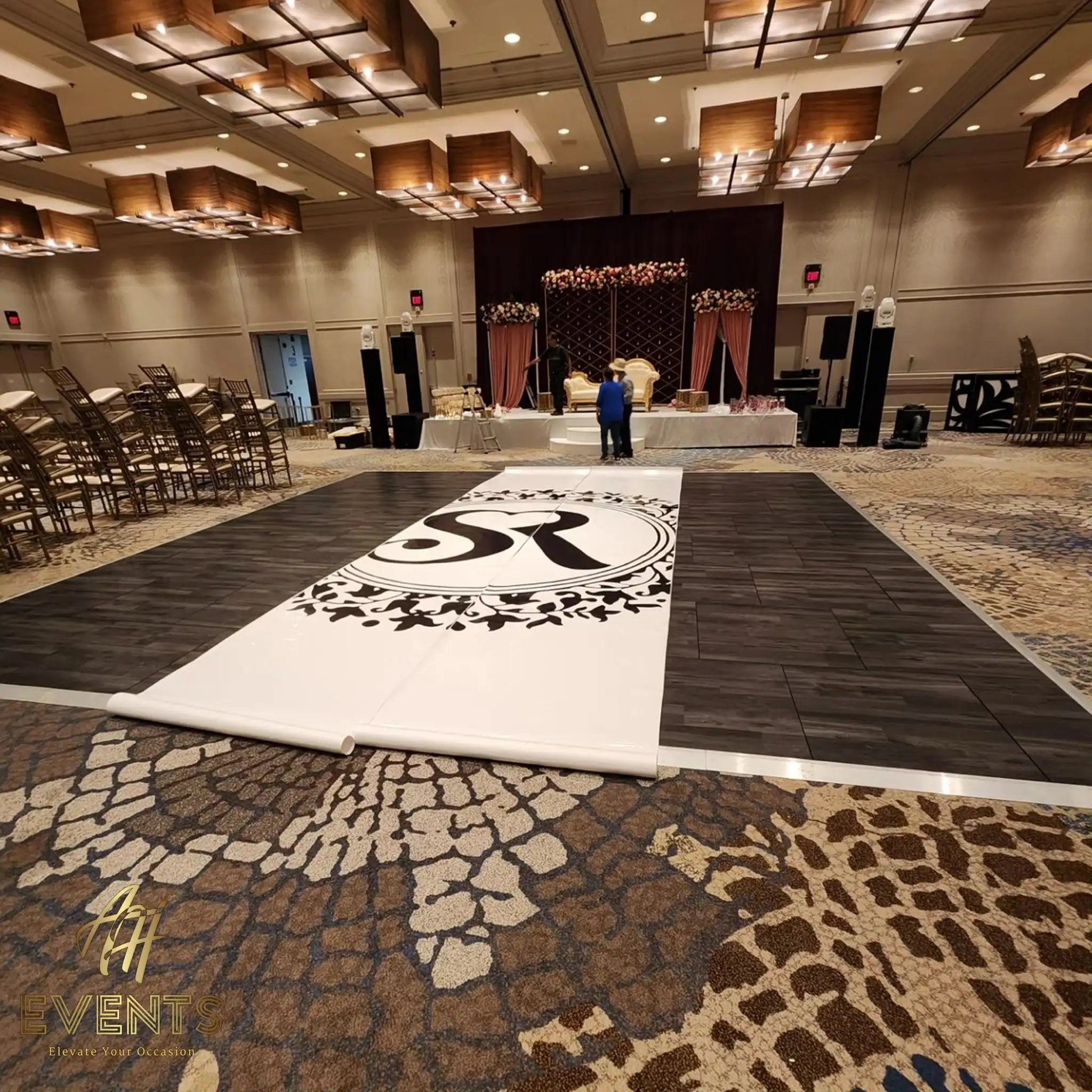 White 'SR' monogram dance floor partly laid over dark tile during setup, the mauve floral stage behind