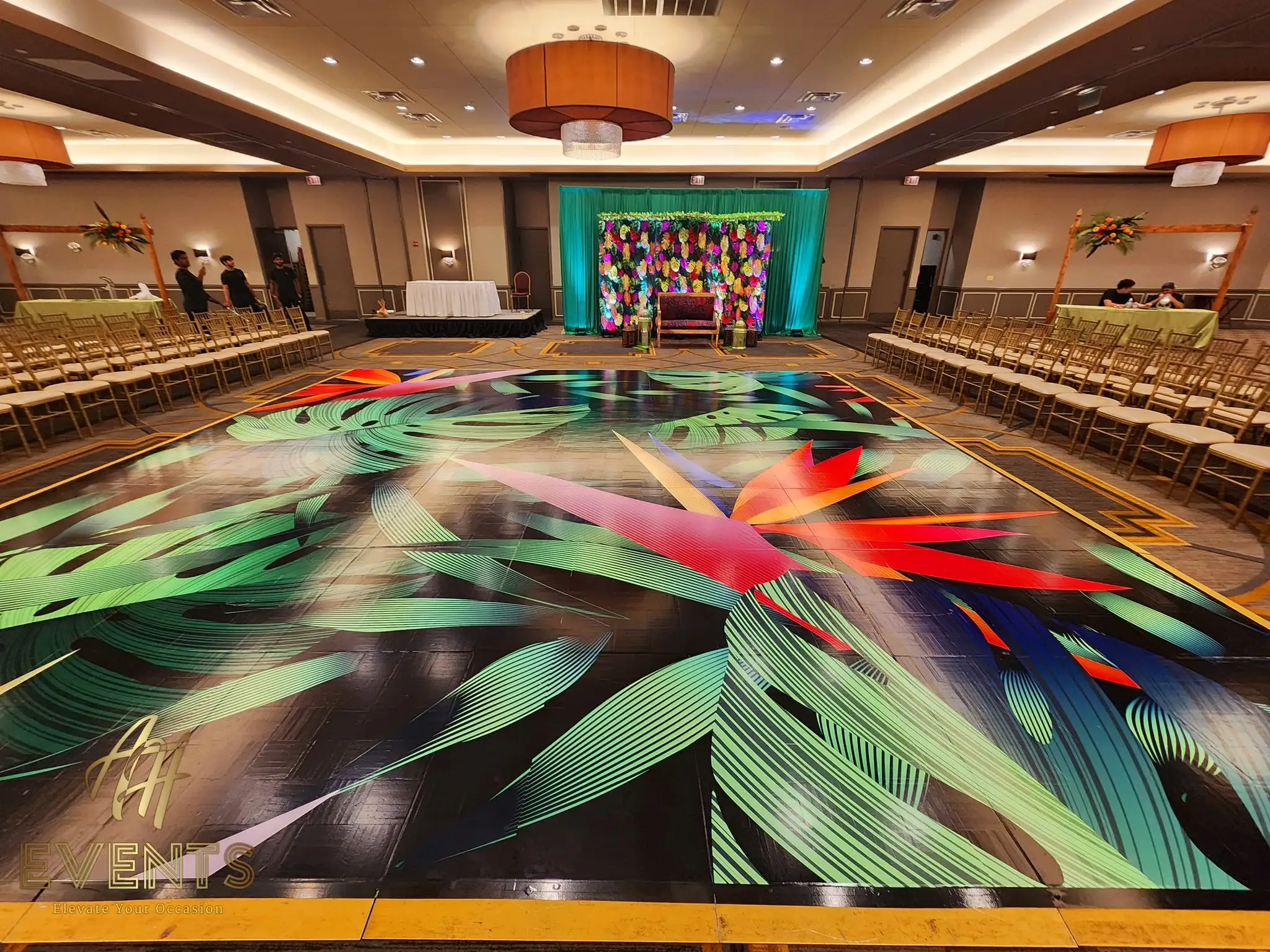 Large dance floor printed with vivid tropical leaf art on black, facing a teal-draped floral stage in a ballroom
