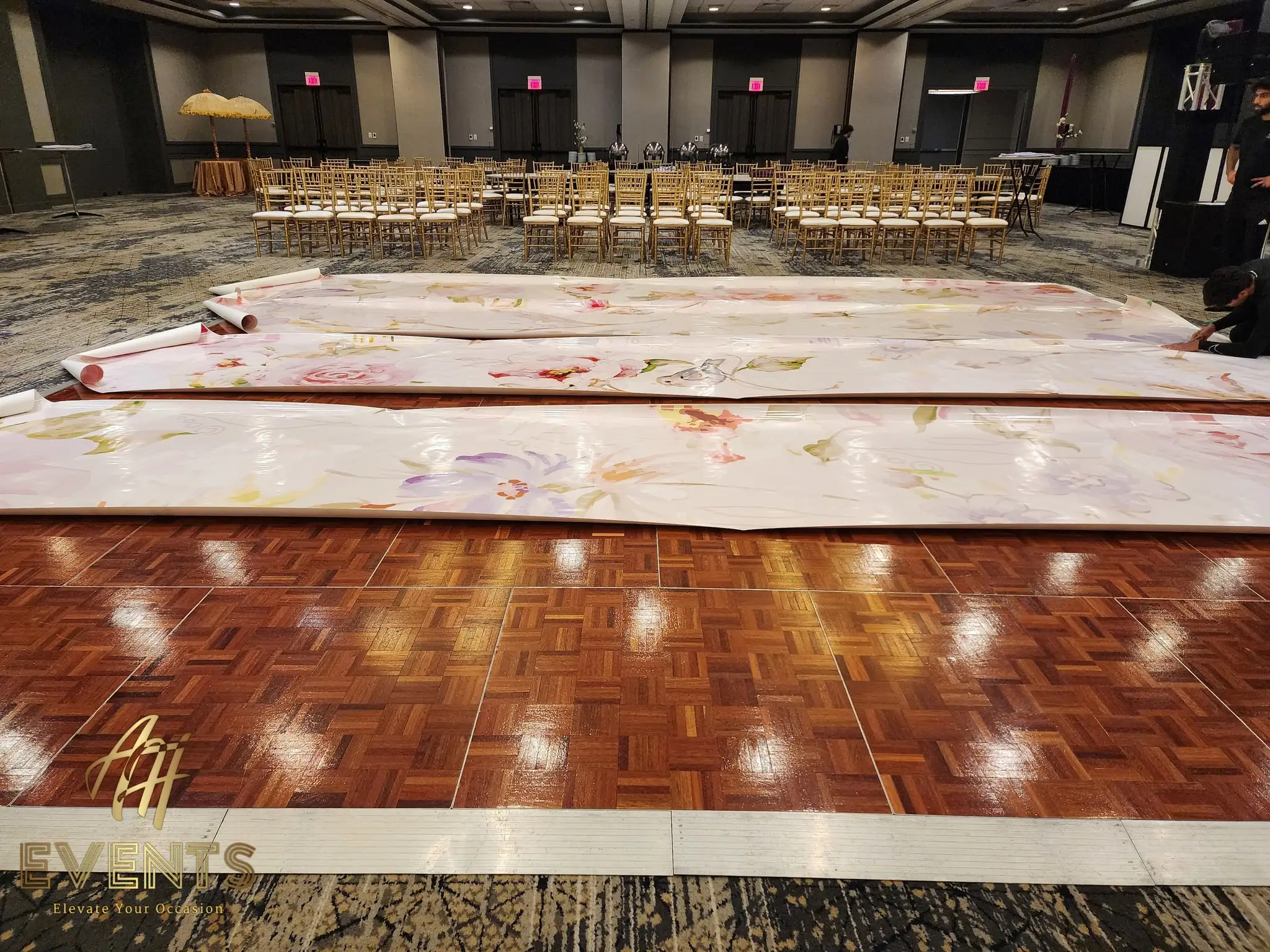 Rolls of the pastel floral dance floor being unrolled over parquet during setup in a ballroom