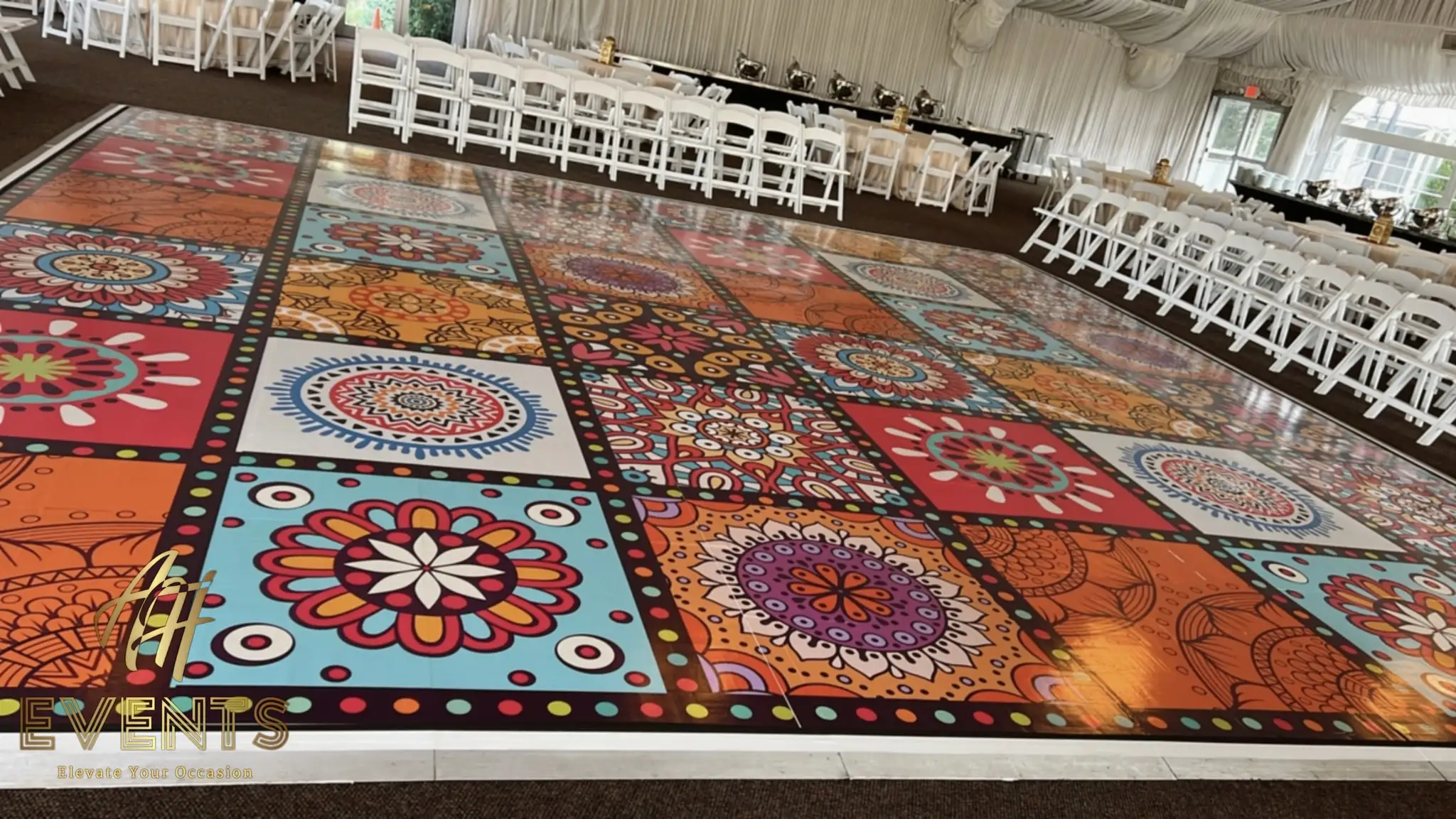 Mandala-tiled dance floor in warm jewel tones under a draped tent, white chairs and tables around