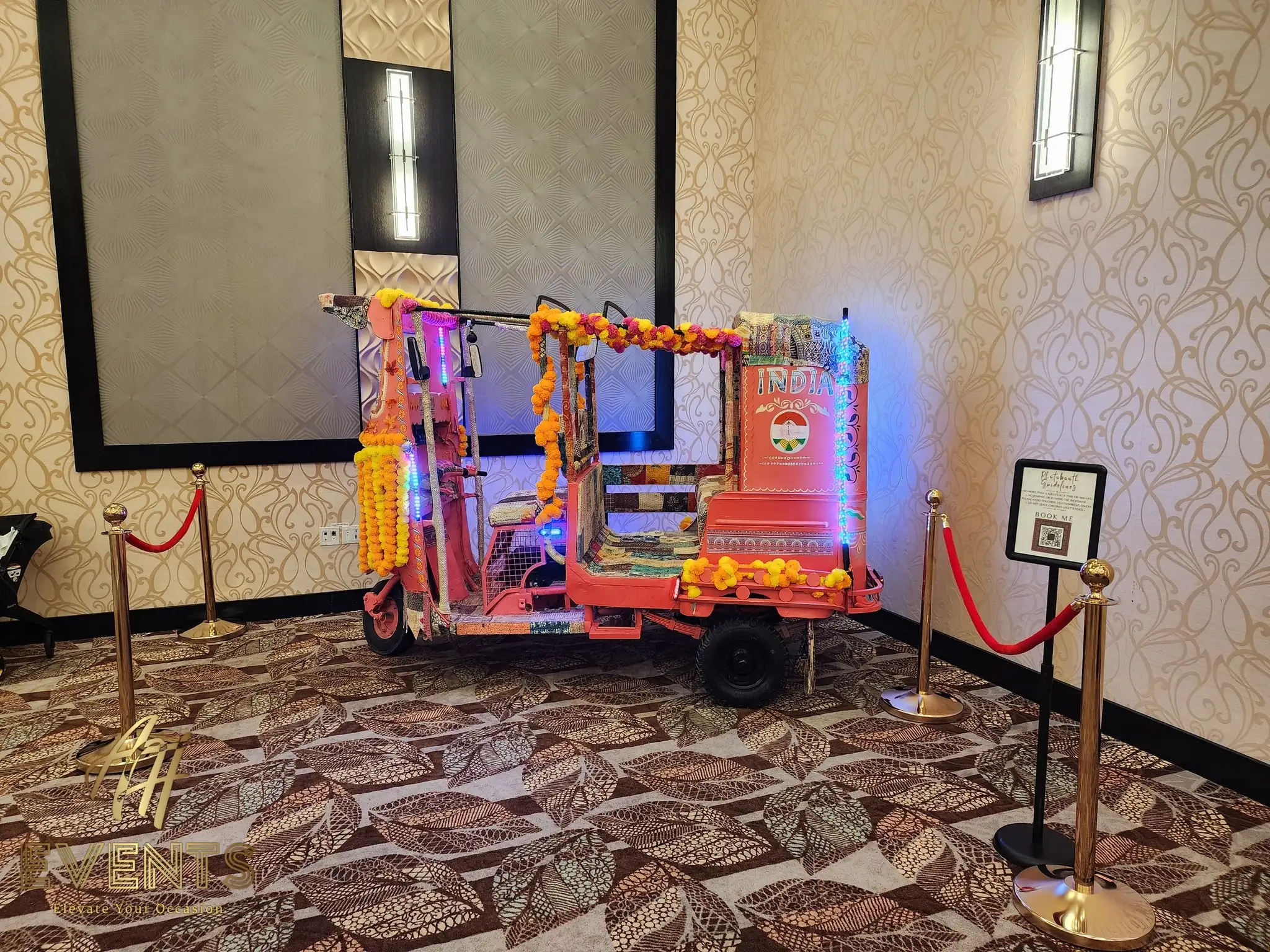 Decorated auto rickshaw with marigolds and LED lighting on display in a hall beside a velvet rope and signage