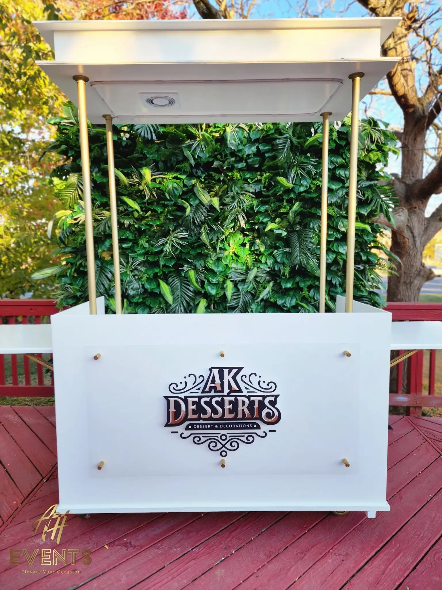White AK Desserts cart with brass canopy poles and a backlit logo panel, set against a green foliage wall outdoors