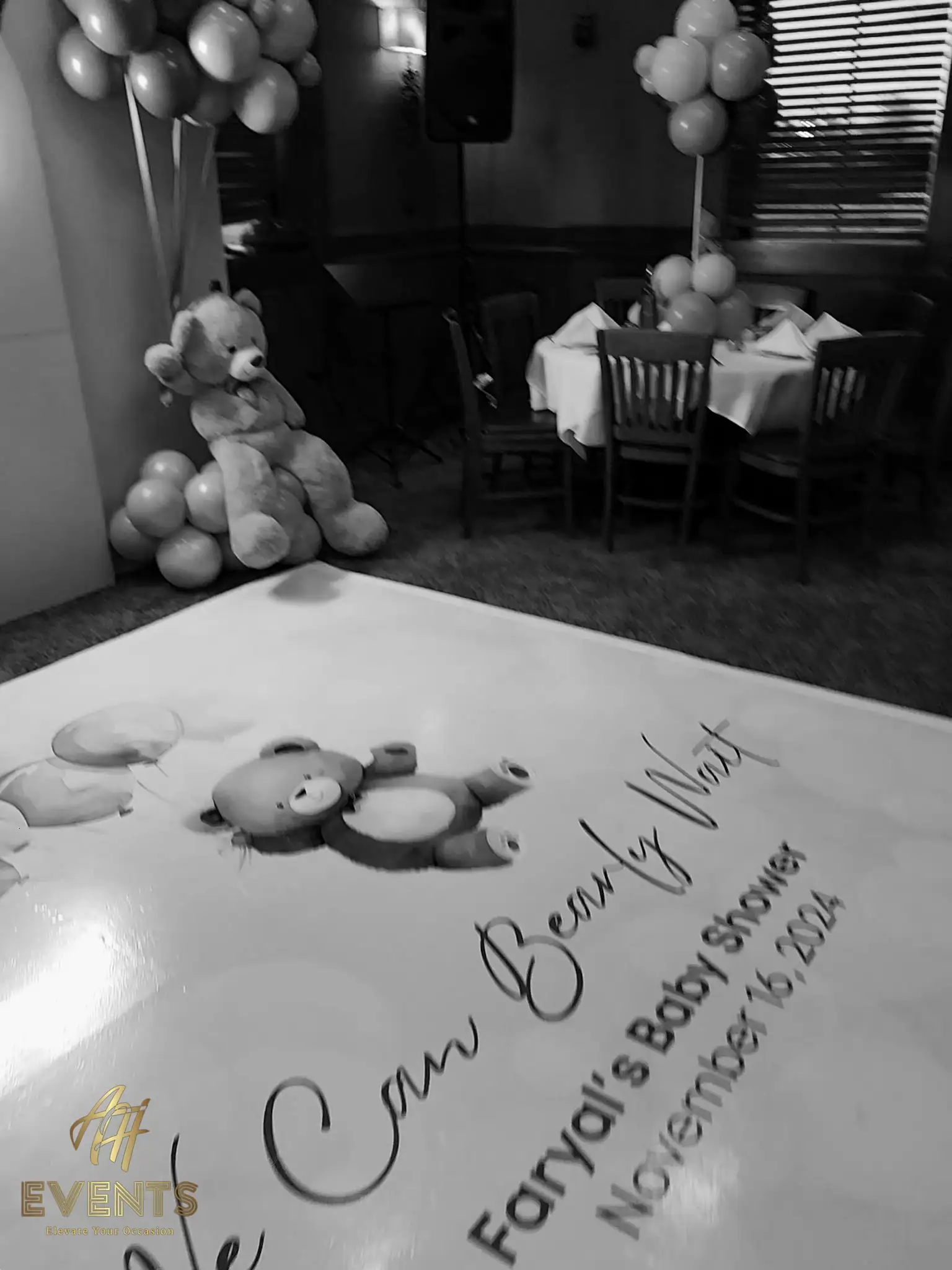 Black-and-white view of the 'We Can Bearly Wait' bear baby shower floor decal, a giant teddy bear and set tables behind