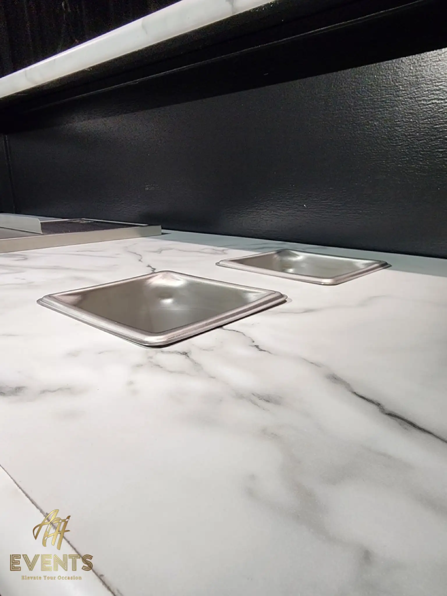 Two recessed stainless condiment pans set into the white marble countertop of the Bobawala cart, black backsplash behind