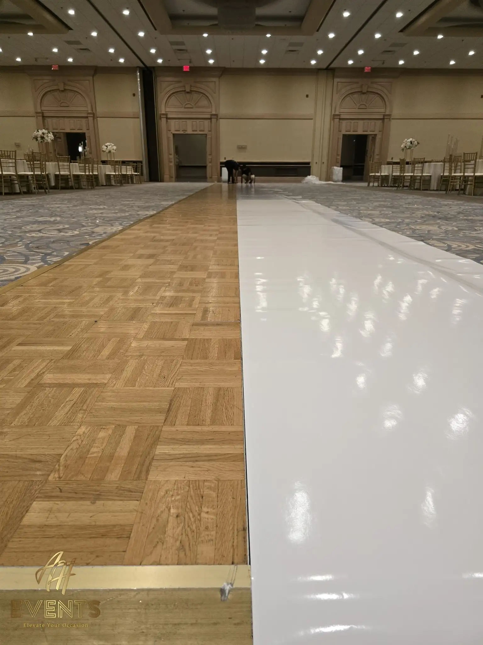 Glossy white vinyl aisle runner laid over a parquet floor during setup in an empty ballroom