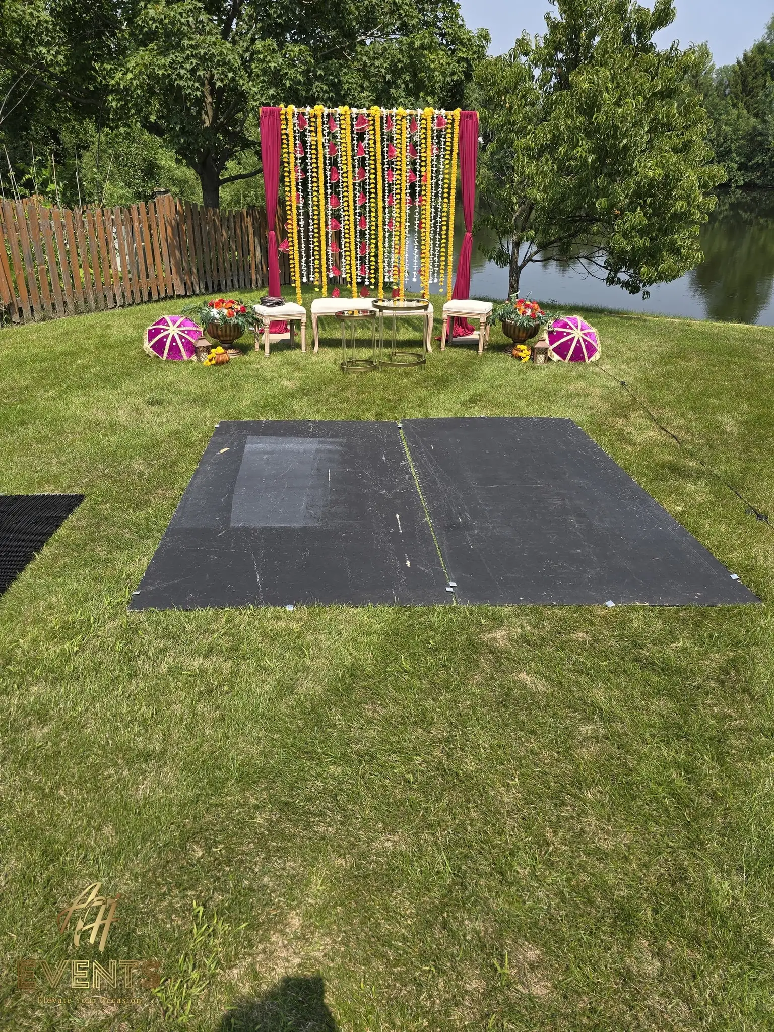 Black subfloor platform on a lawn by a pond with a marigold-curtain backdrop, before the mandala floor is laid