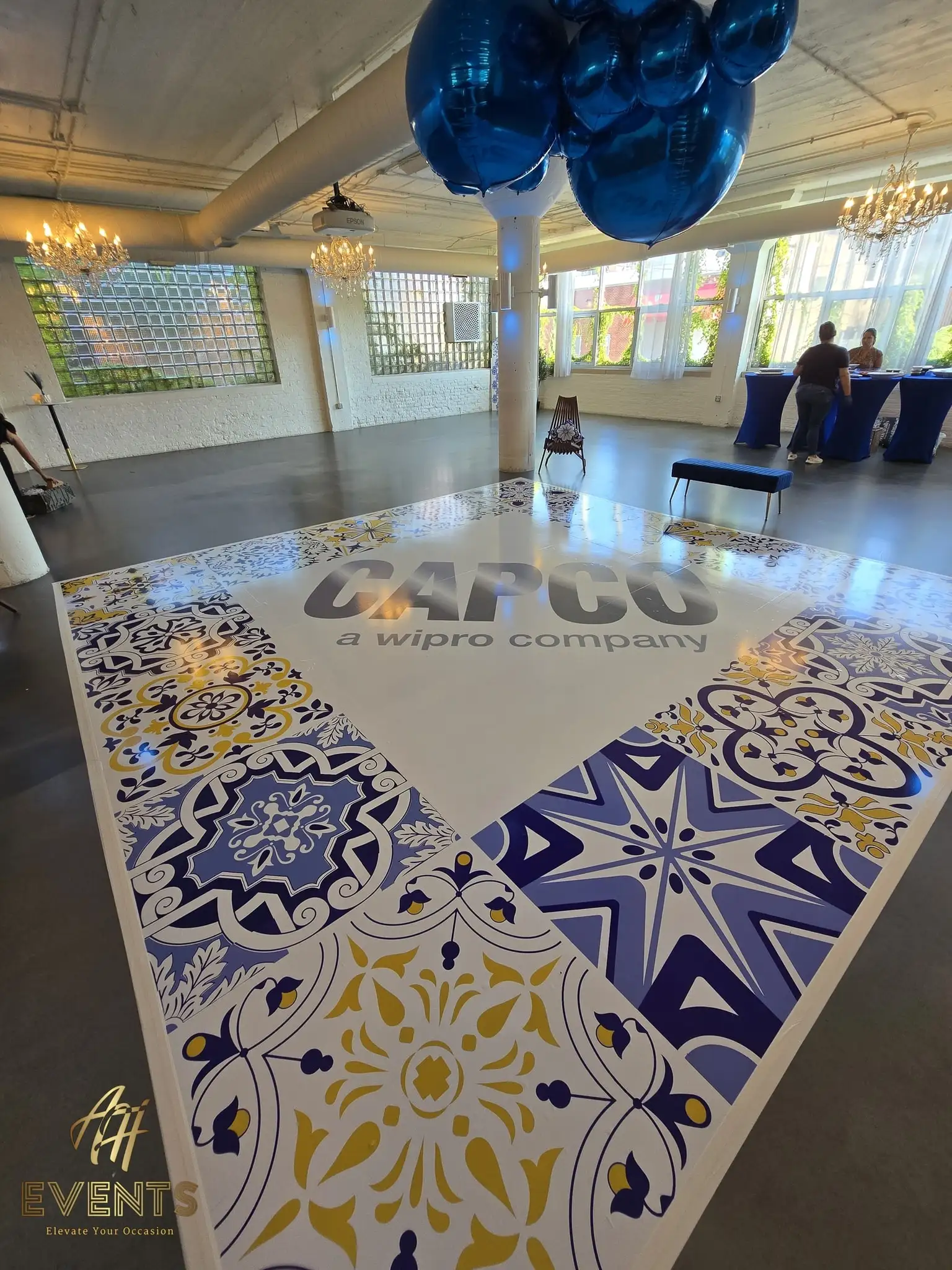 Corporate dance floor with a blue-and-yellow tile border and the CAPCO logo in a loft venue, blue balloons overhead