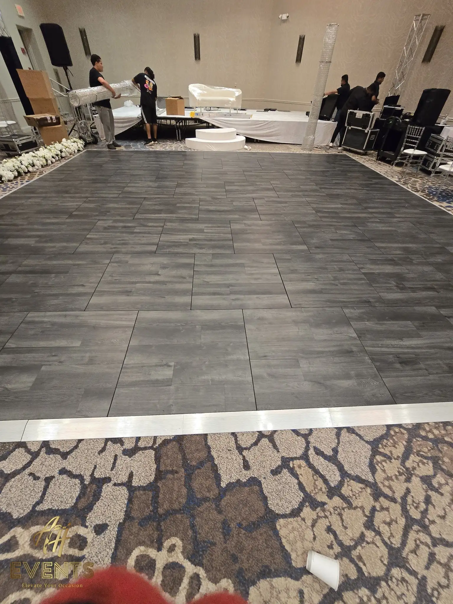 Charcoal-gray plank dance floor being installed in a ballroom during setup, white vinyl rolls nearby