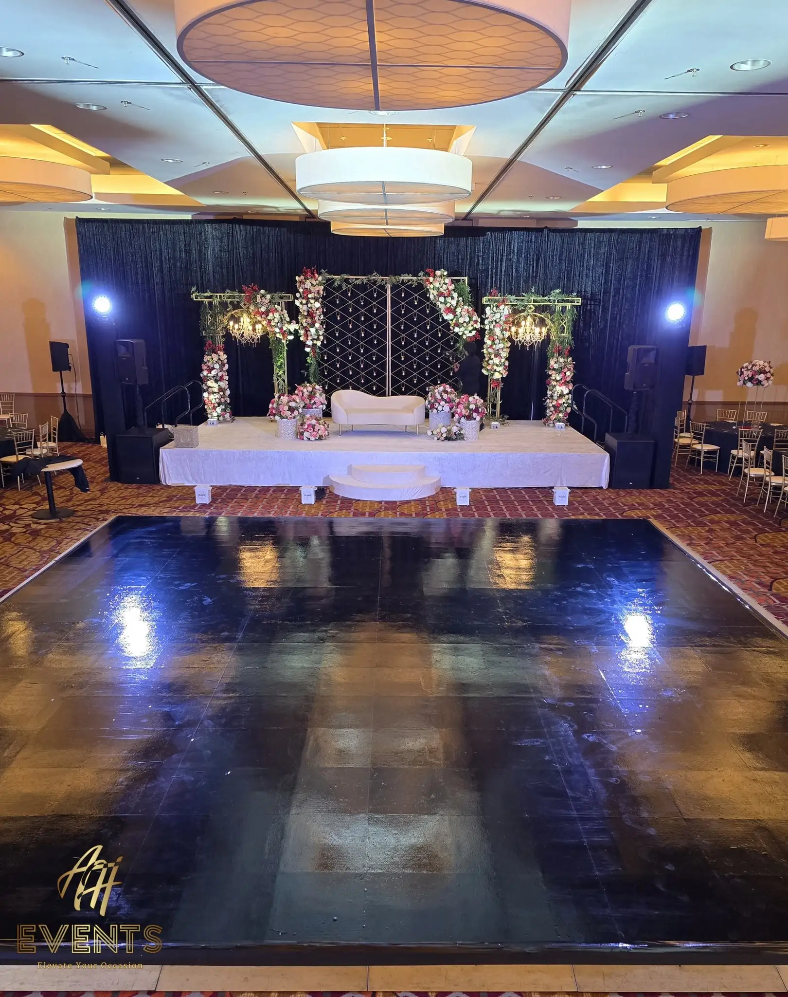 Black dance floor before a white stage with a floral mandap and gold lattice screens under a navy drape
