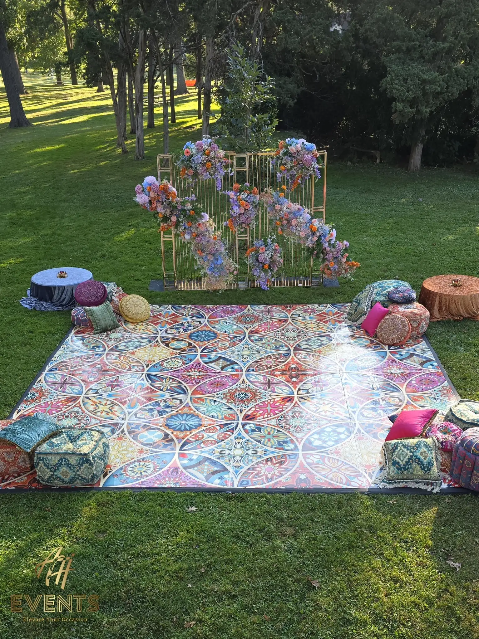 Outdoor Kaleidoscope floor with a gold floral-draped arch and patterned floor cushions on a sunny lawn