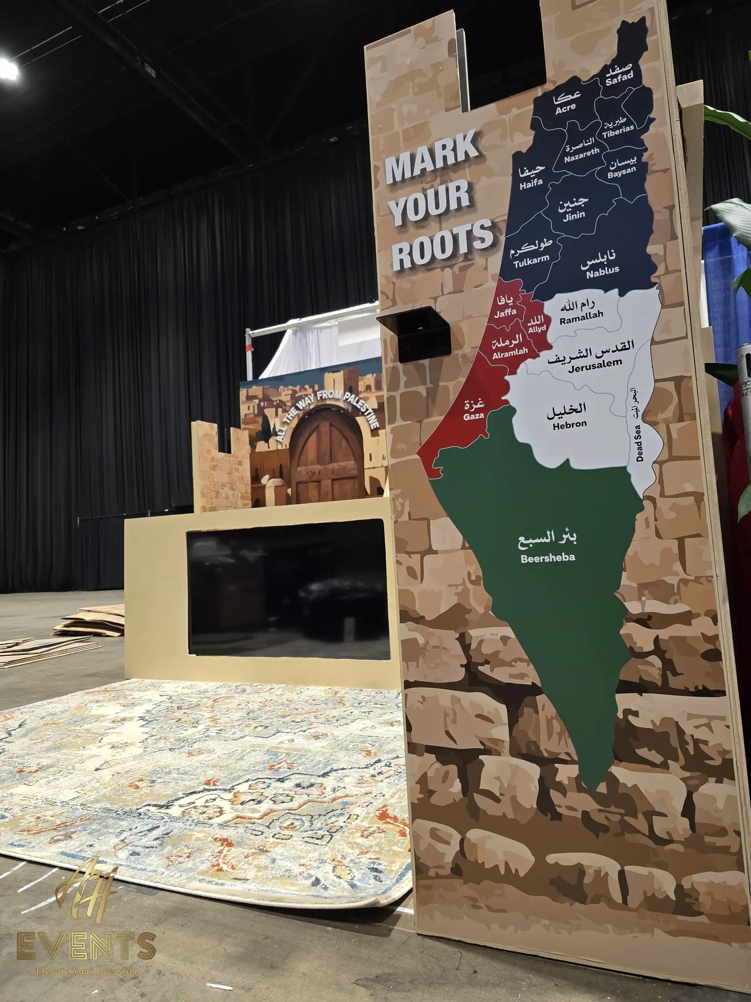 'Mark Your Roots' booth panel with a map of Palestine beside a brick-print wall and arched door