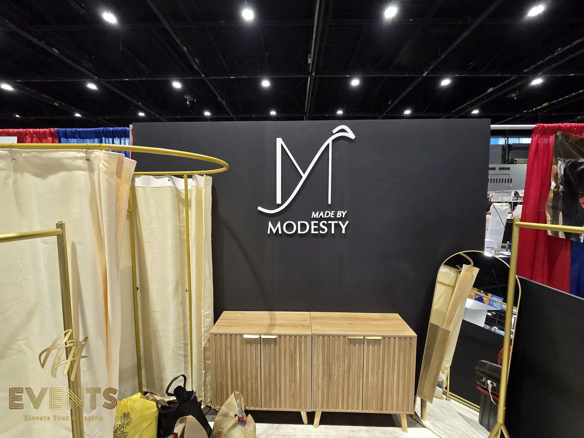 Made by Modesty booth showing the logo wall, gold garment racks and a wood cabinet on the convention floor