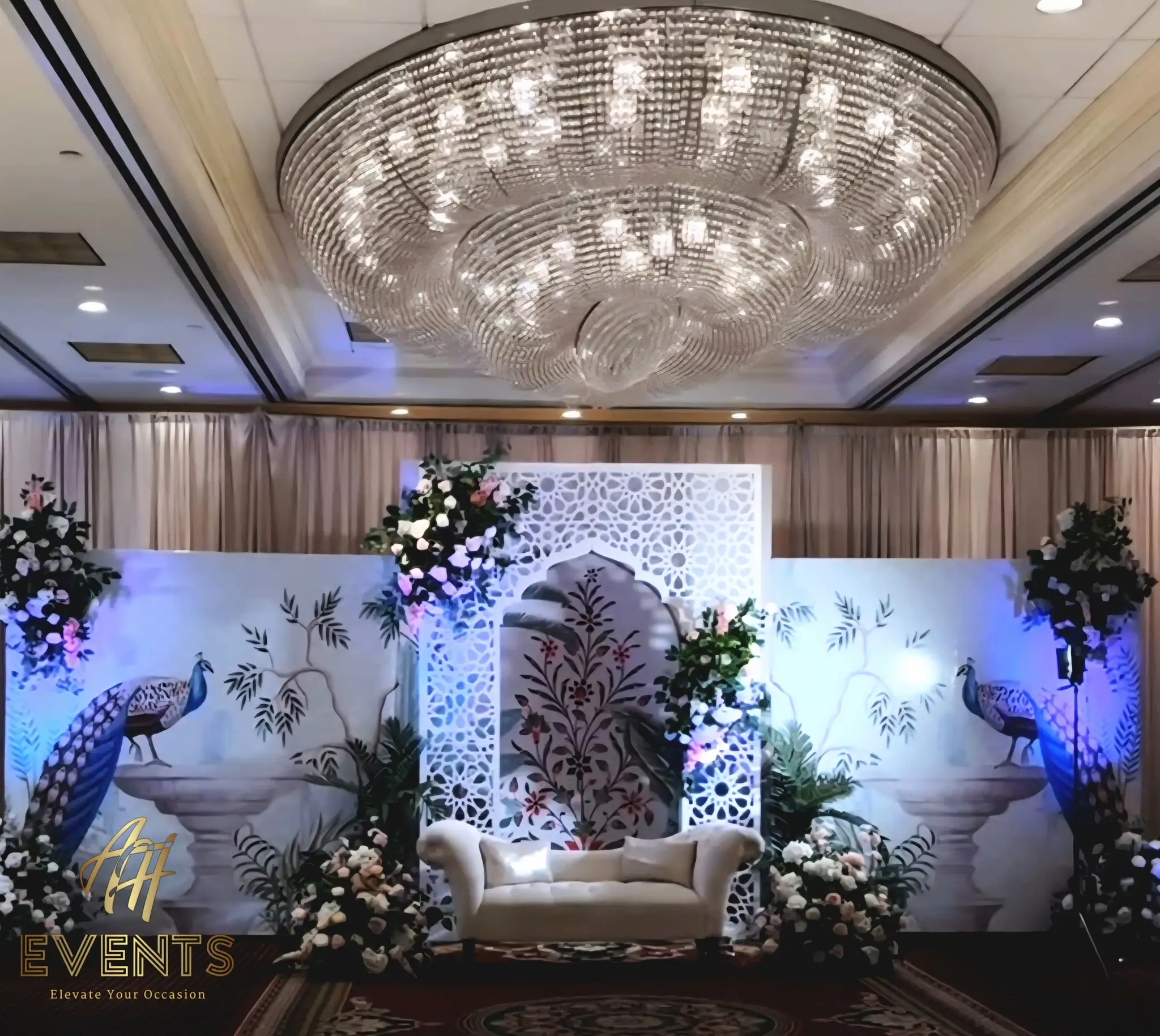 White matrix arch on a ballroom stage flanked by peacock-print panels and florals under a large chandelier
