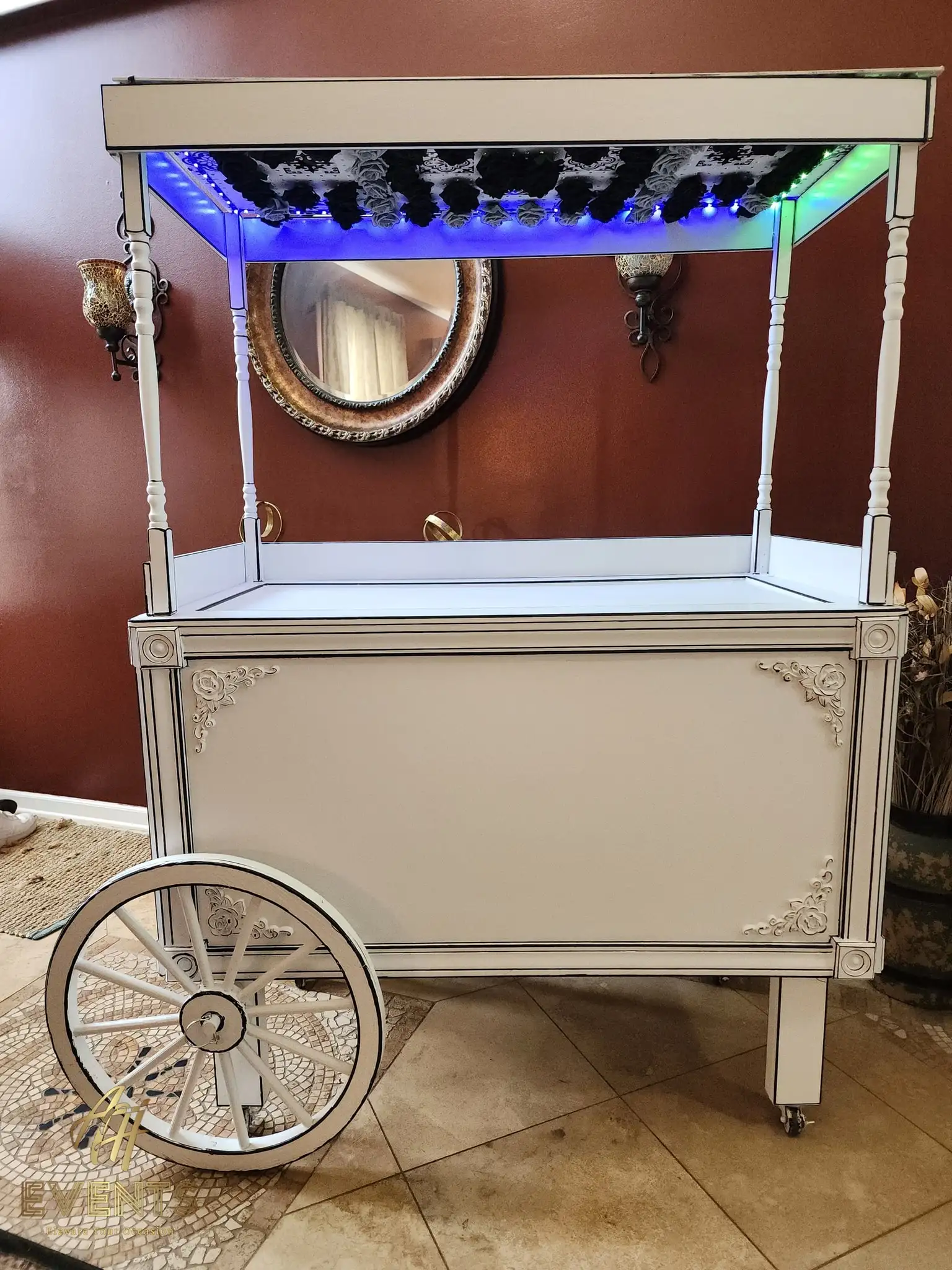 White display cart with an LED-lit canopy and decorative scrollwork, set against a round gold mirror