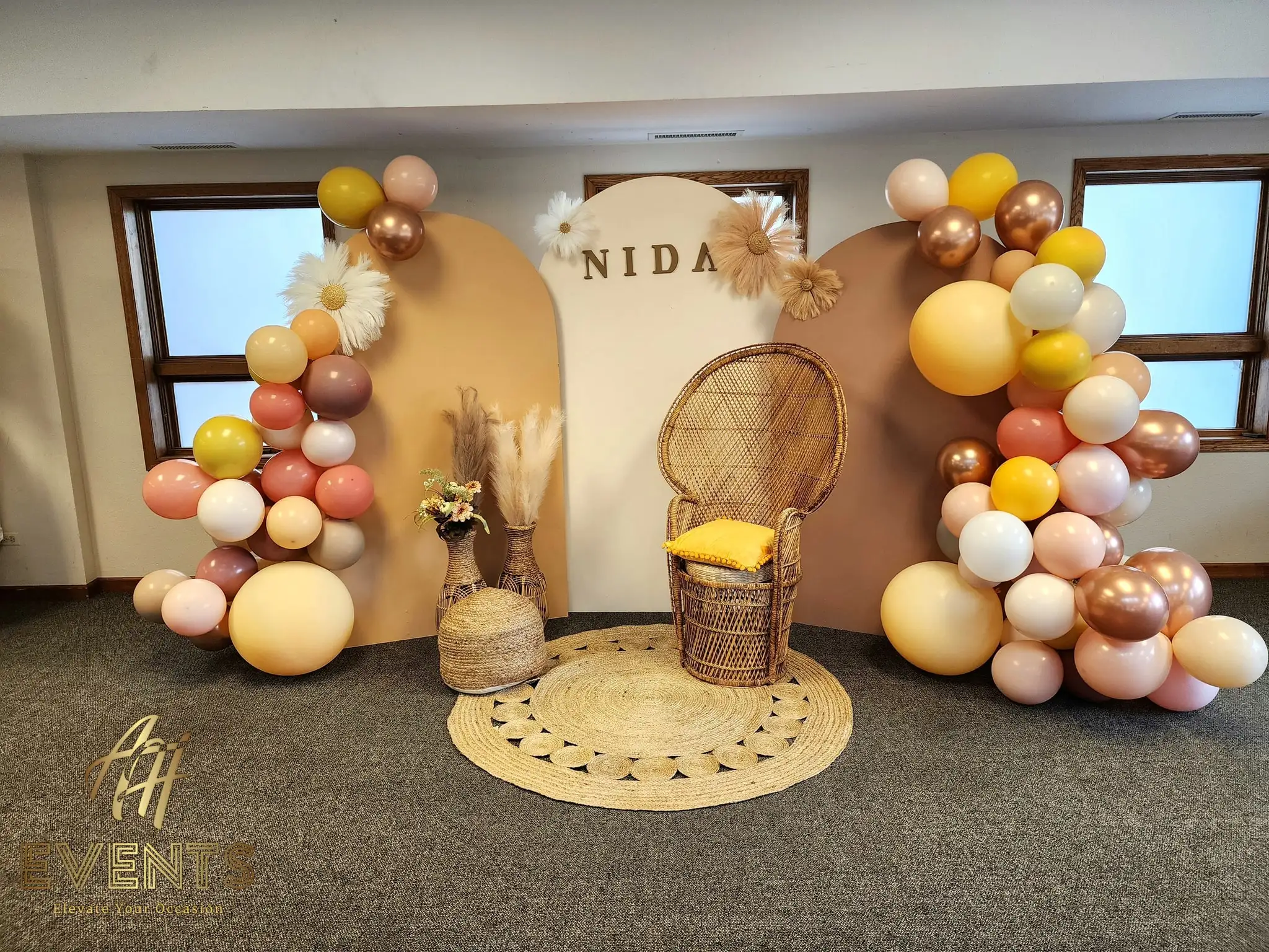 Boho 'Nida' arch backdrop flanked by mustard and cream balloon garlands with a peacock chair on a jute rug