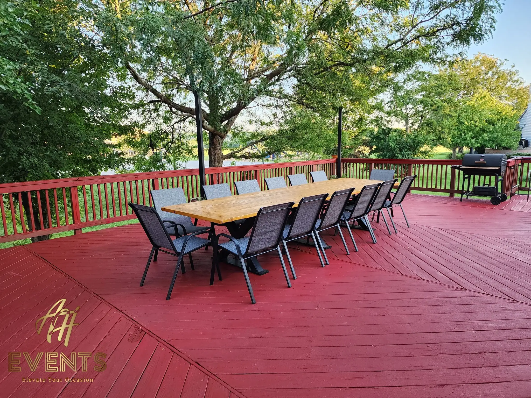 Wooden farm table and gray chairs on a red deck, a grill and lawn in the background