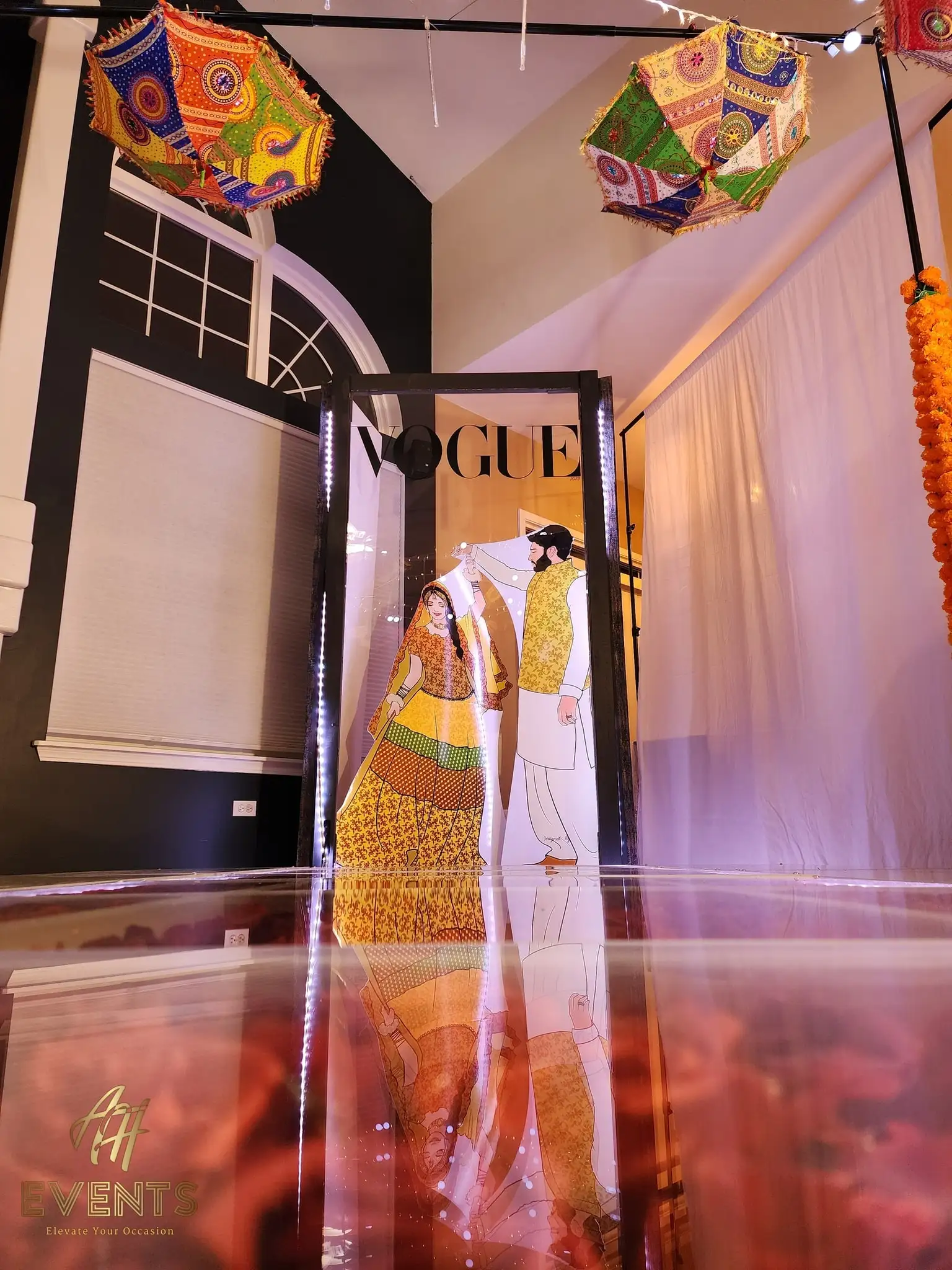 Couple posing in a VOGUE-branded mirror photo booth with colorful umbrellas overhead and a red-rose glass floor
