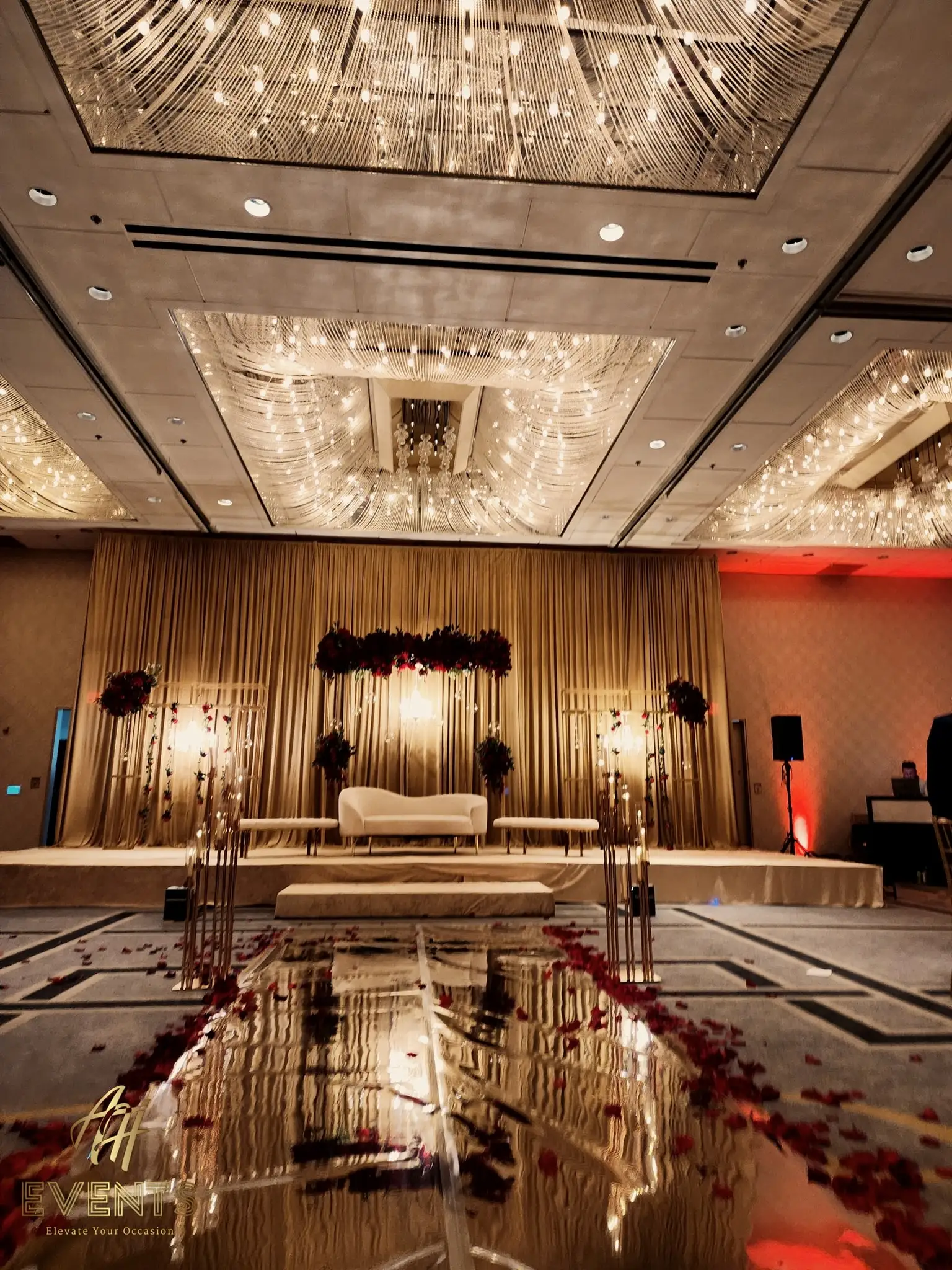 Gold-draped wedding stage with a white sofa at the end of a reflective mirror vinyl aisle scattered with red rose petals