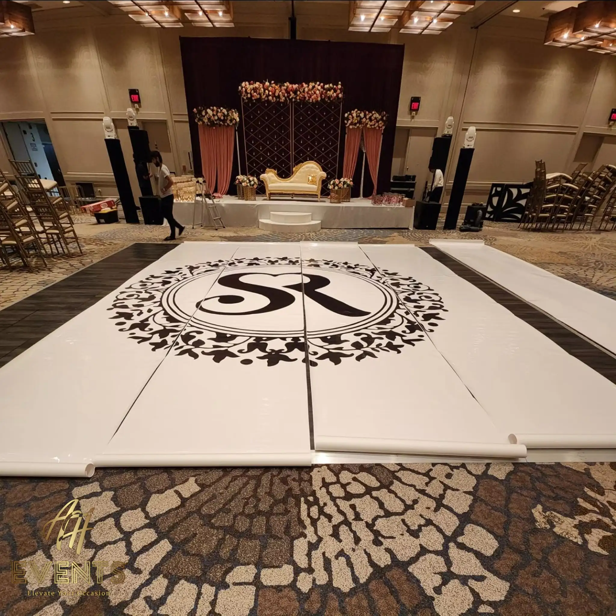 White dance floor with the 'SR' floral-roundel monogram, a gold sofa stage behind, one section still being unrolled