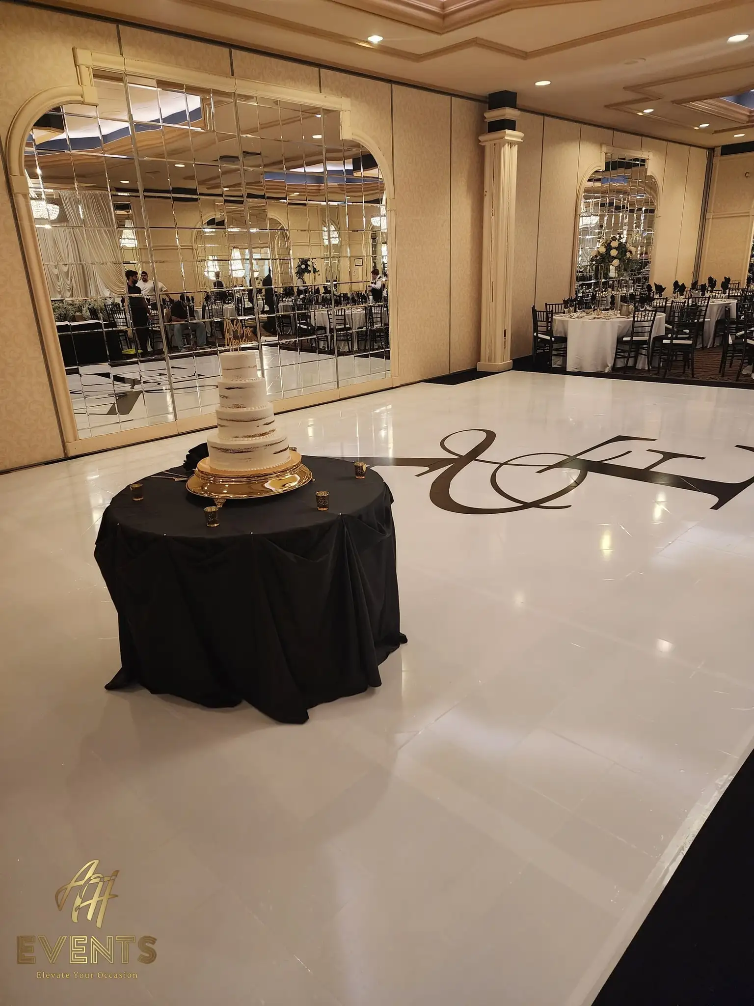 Tiered wedding cake on a black-draped round table beside the white 'A&E' monogram dance floor and mirrored wall