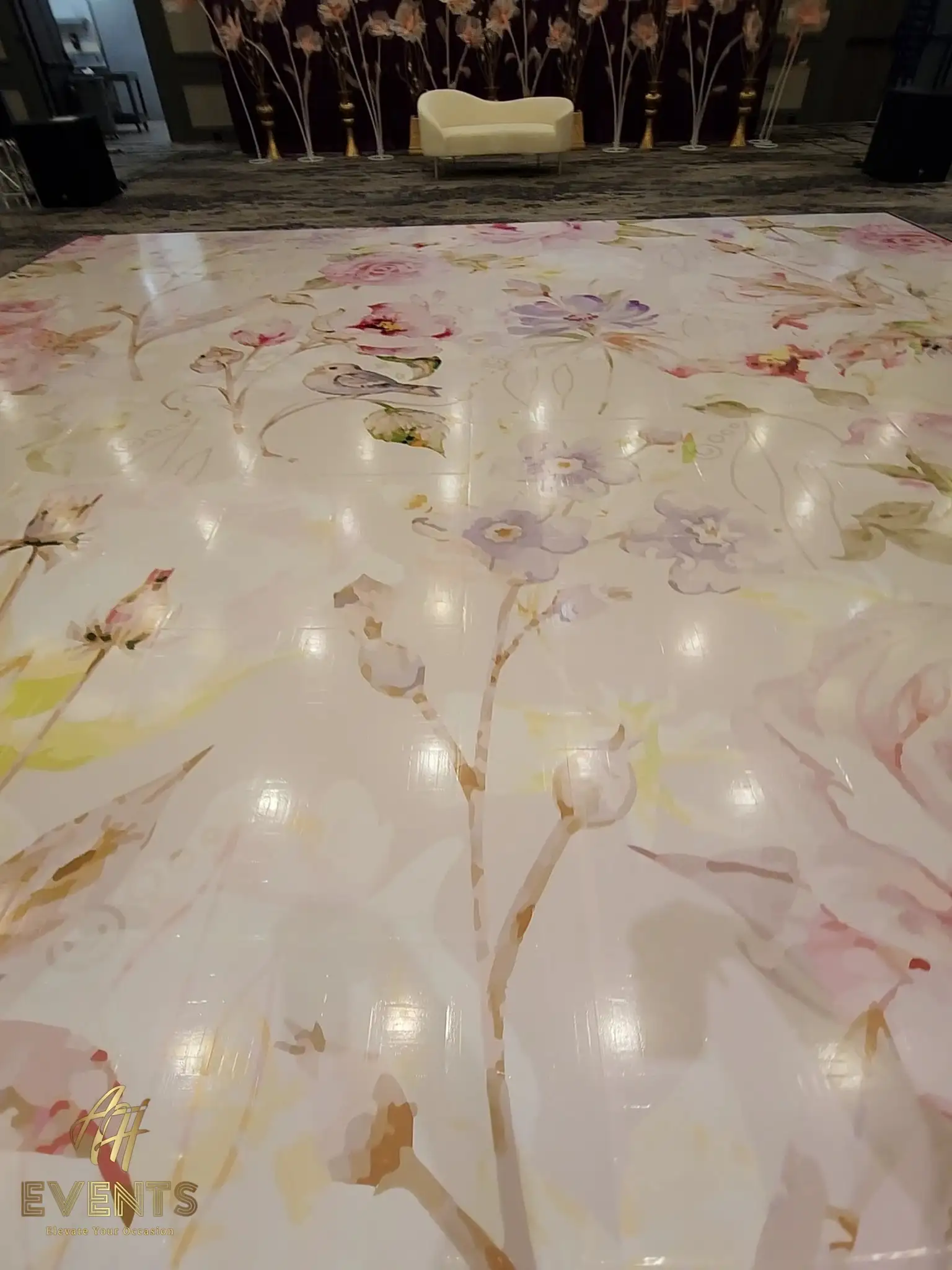 Pastel floral dance floor facing a stage with tall floral stands and a white sofa