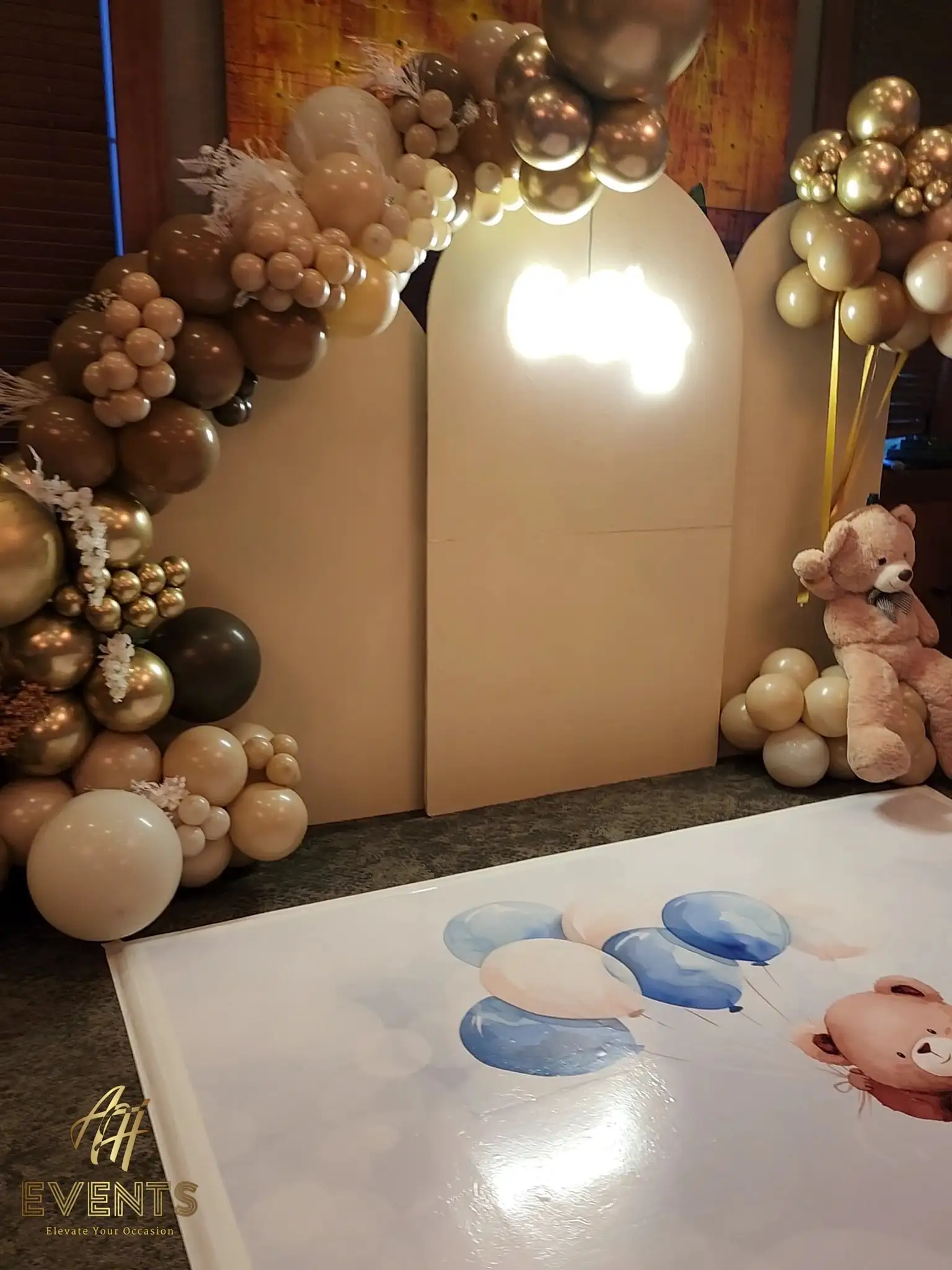 Bear baby shower backdrop of tan arches and balloon garlands beside the printed floor decal showing a teddy holding blue and cream balloons