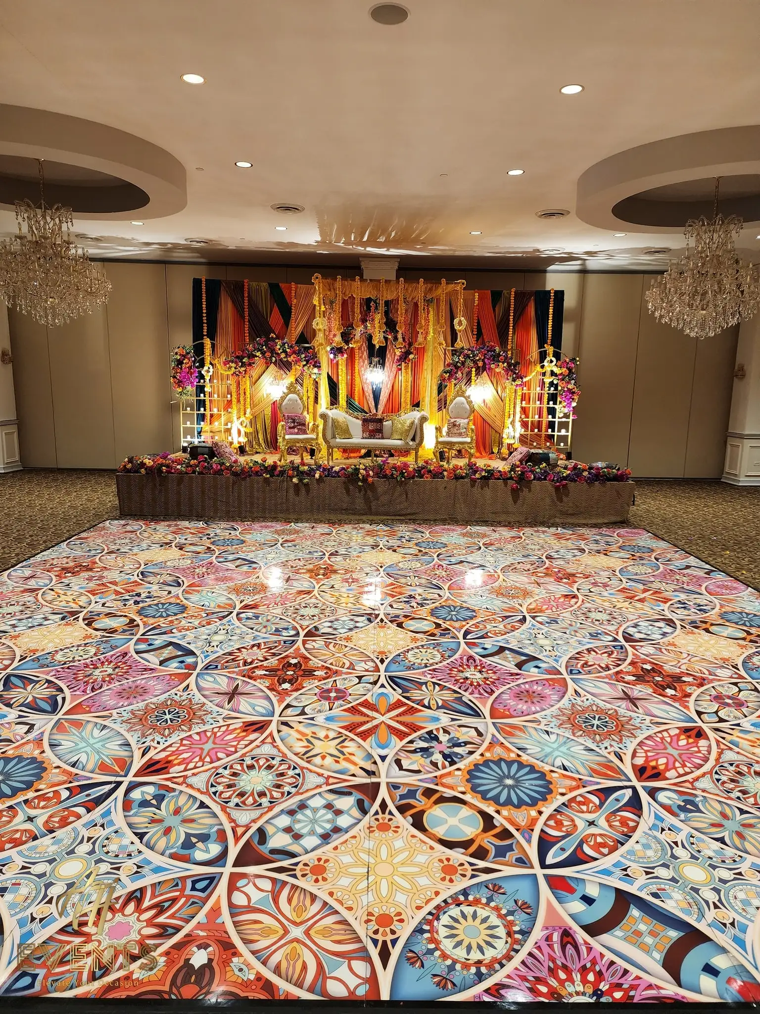 Colorful Kaleidoscope floor in front of a gold-and-orange draped wedding stage with thrones, flanked by chandeliers