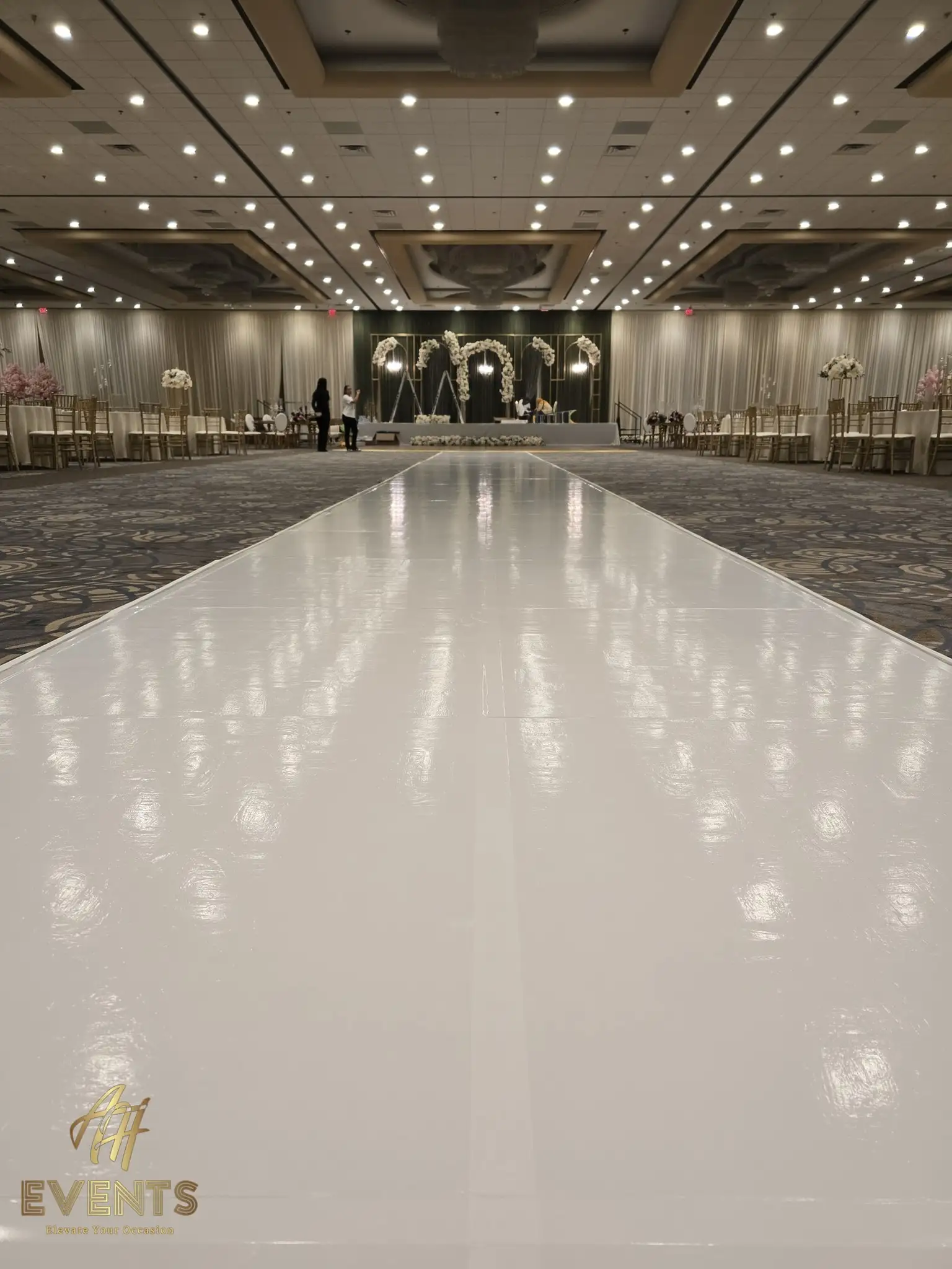 Glossy white vinyl runway leading to a white-floral arch stage, lined with gold chiavari chairs
