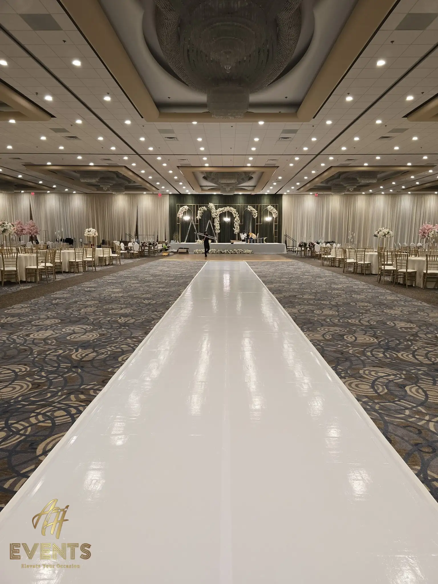 Glossy white vinyl runway running the length of a ballroom to a floral arch stage beneath an ornate ceiling