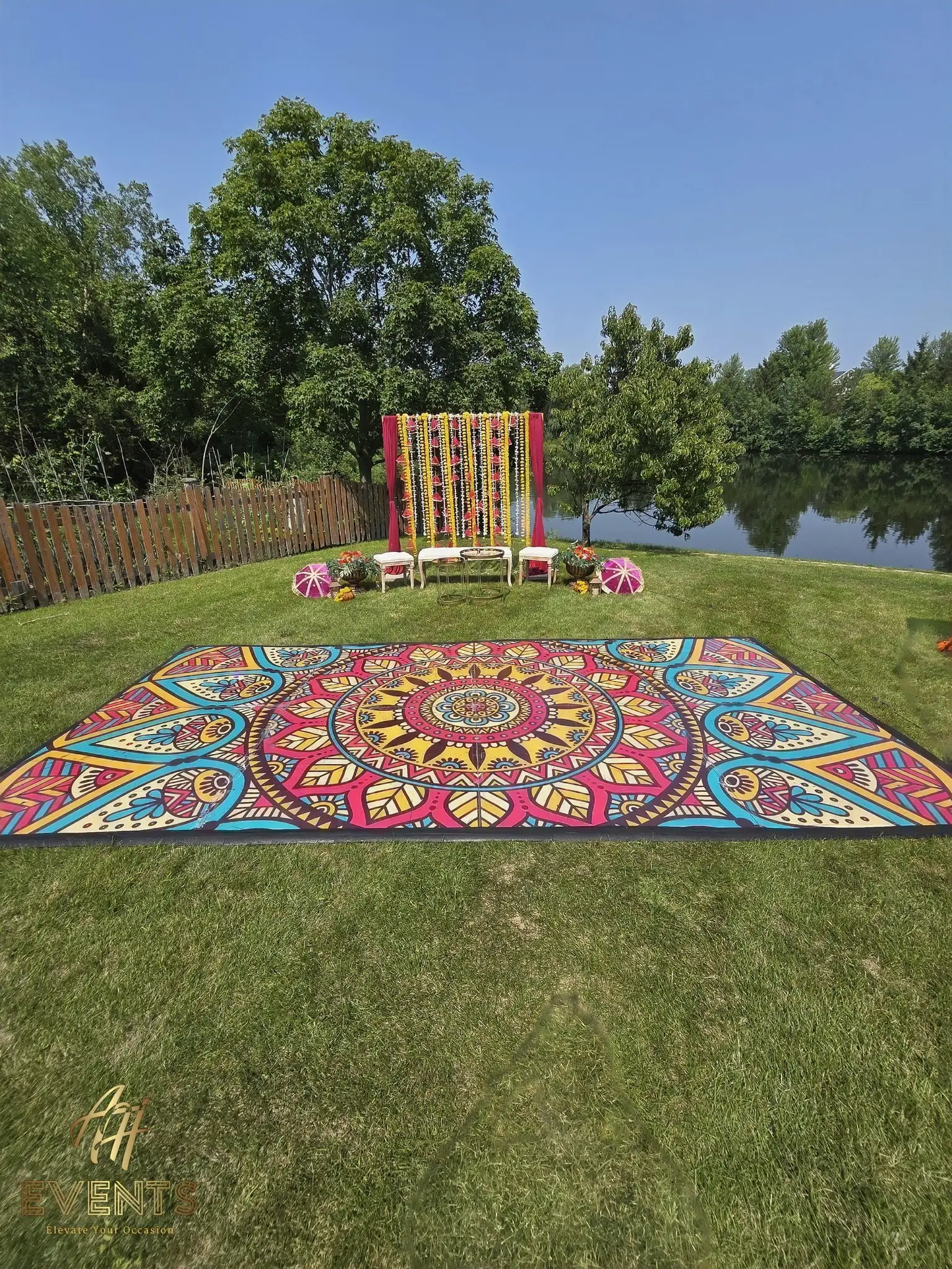 Mandala floor on a sunny lawn with a pink-and-yellow drape backdrop beside a pond