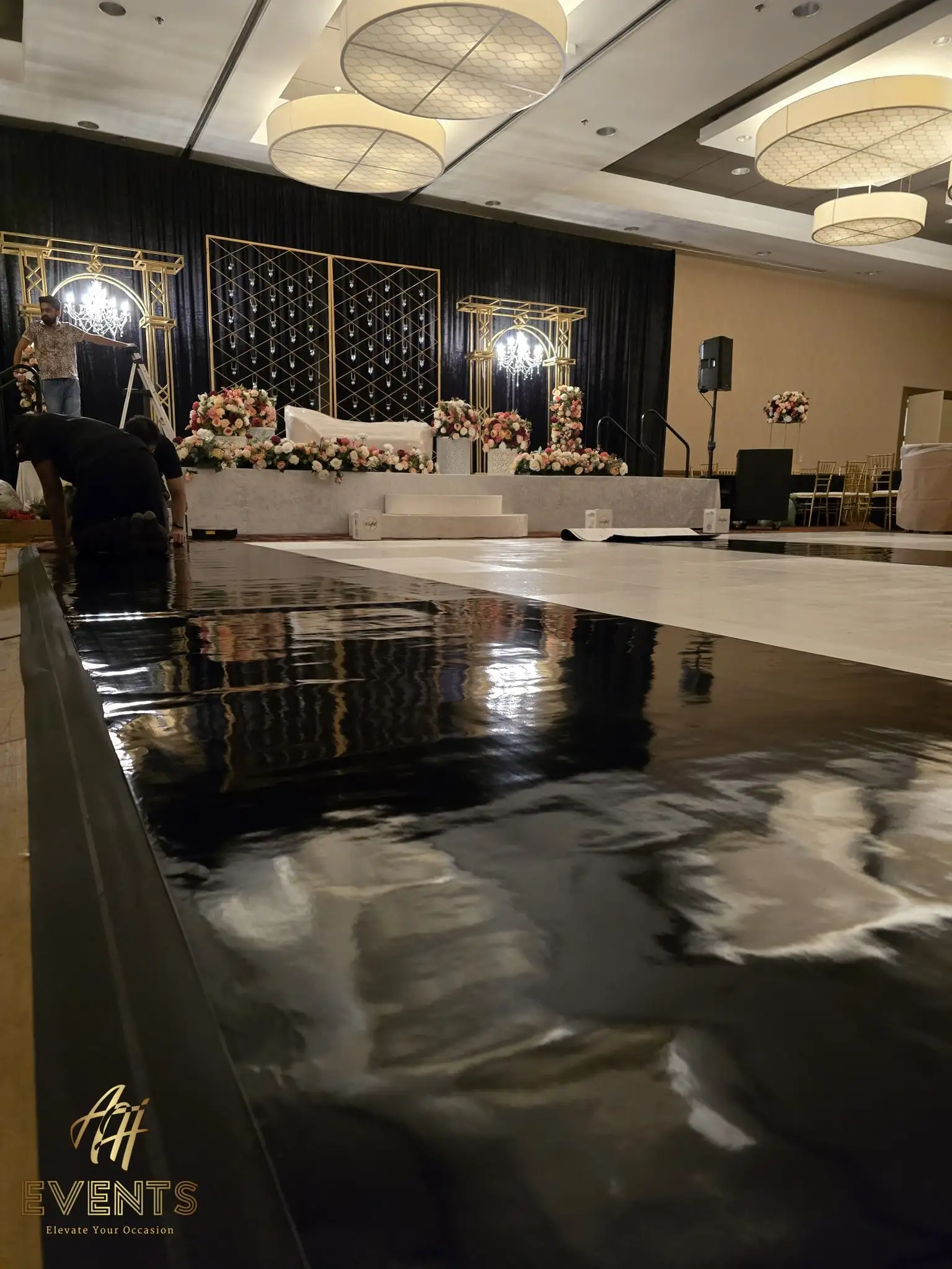 Installers laying the black-and-white dance floor near a flower-trimmed gold-framed stage during setup